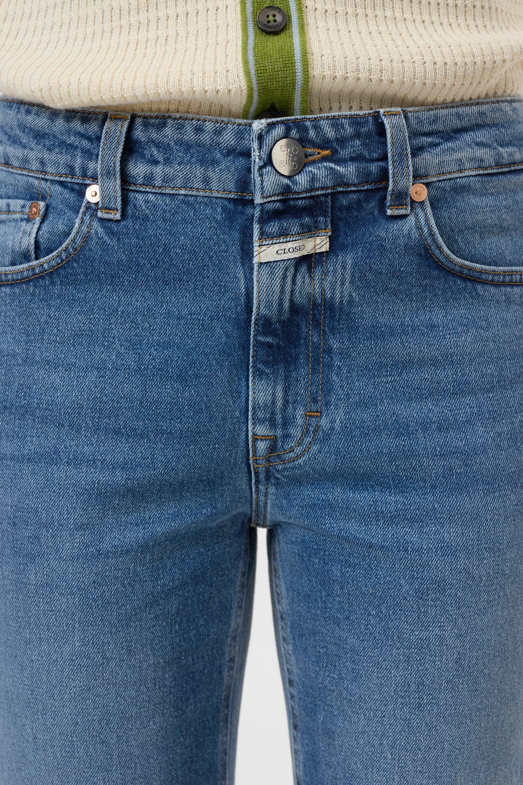 Close-up of a person wearing CLOSED's Milo straight-leg jeans made from sustainable denim, styled with a cream ribbed sweater featuring green trim and buttons. The jeans are fastened, showing a white branded tag near the zipper.