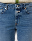 Close-up of a person wearing CLOSED's Milo straight-leg jeans made from sustainable denim, styled with a cream ribbed sweater featuring green trim and buttons. The jeans are fastened, showing a white branded tag near the zipper.