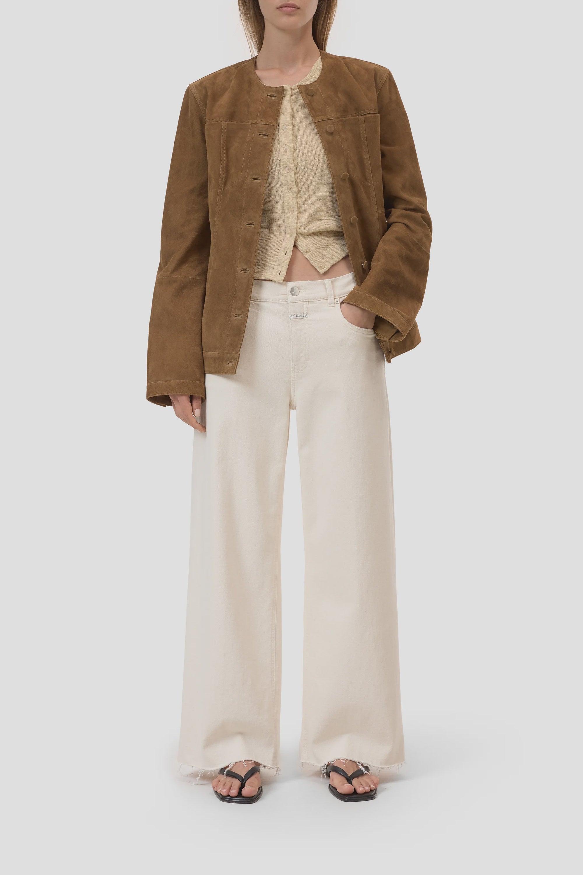 A woman stands against a plain light background wearing CLOSED Jayla Denim wide fit jeans, a brown suede jacket over a beige cardigan, black flip-flop sandals, and her hair is straight.