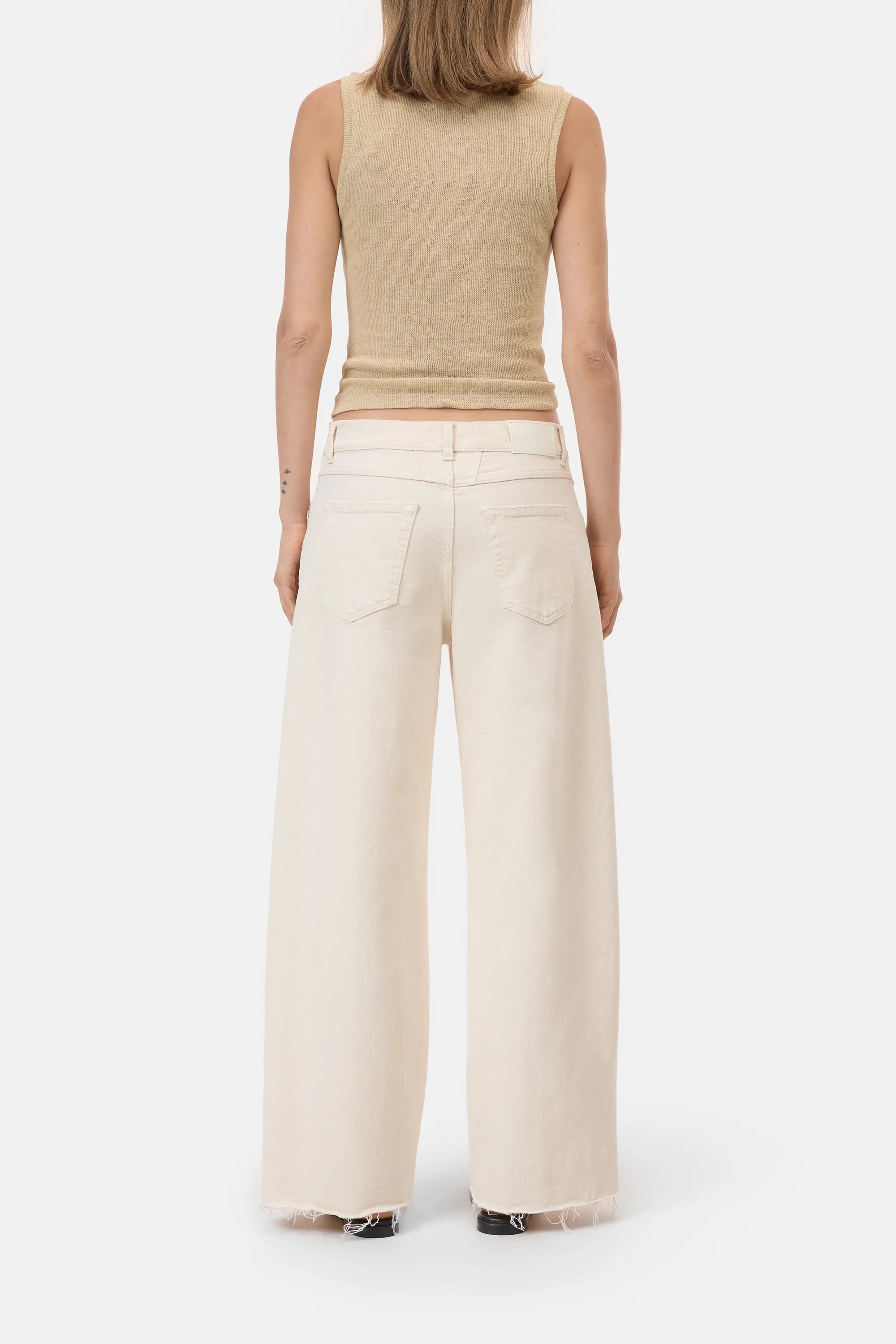 A person stands facing away, wearing CLOSED's Jayla Denim—high-waisted, wide-fit ecru jeans with raw hems and two back pockets, crafted in Italy. The plain white background highlights the beige sleeveless ribbed top.