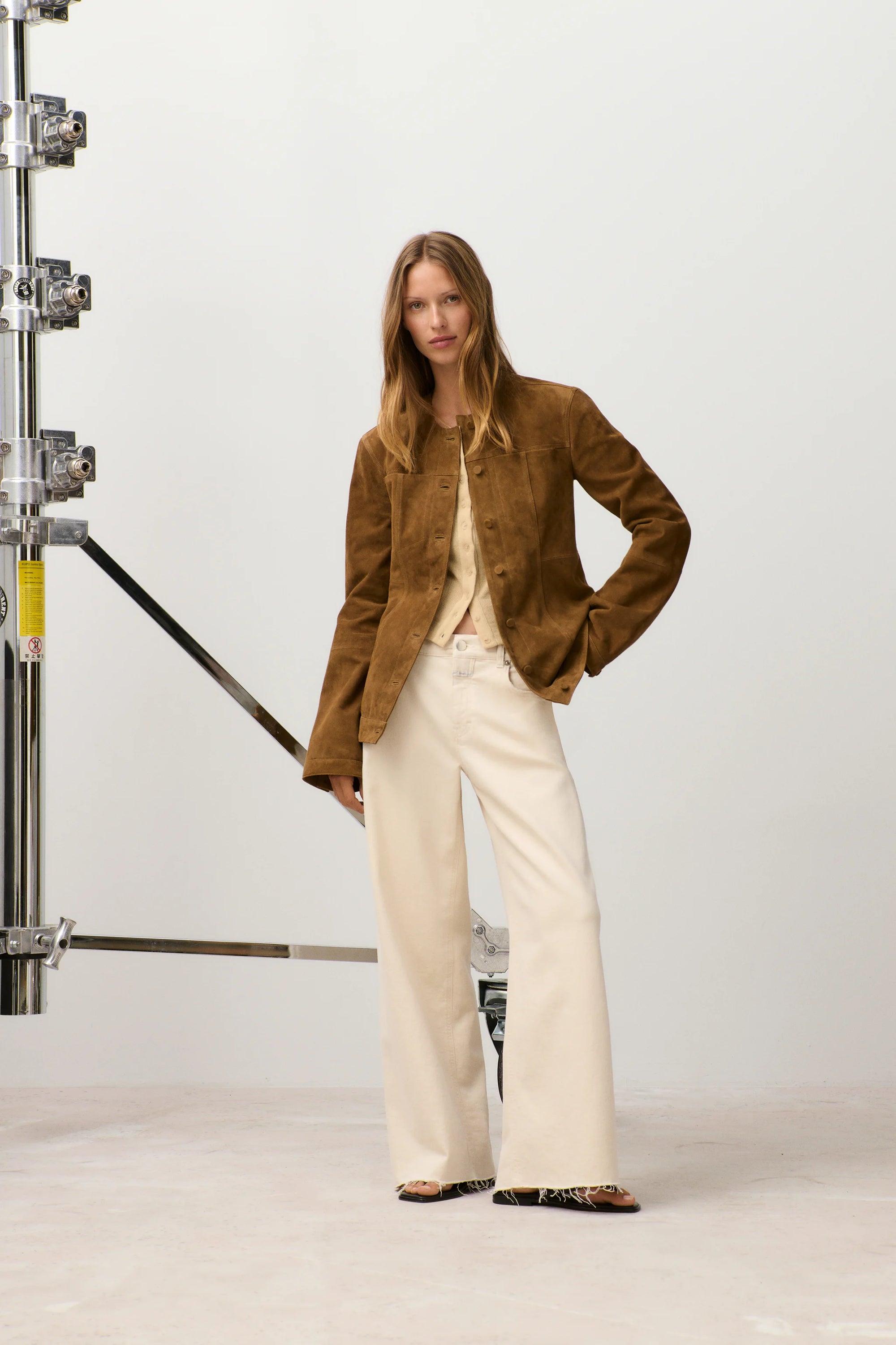 A woman with long, straight hair stands confidently in a studio, wearing the CLOSED Jayla Denim jeans, a brown suede jacket, and a beige top. Industrial equipment can be seen in the background.