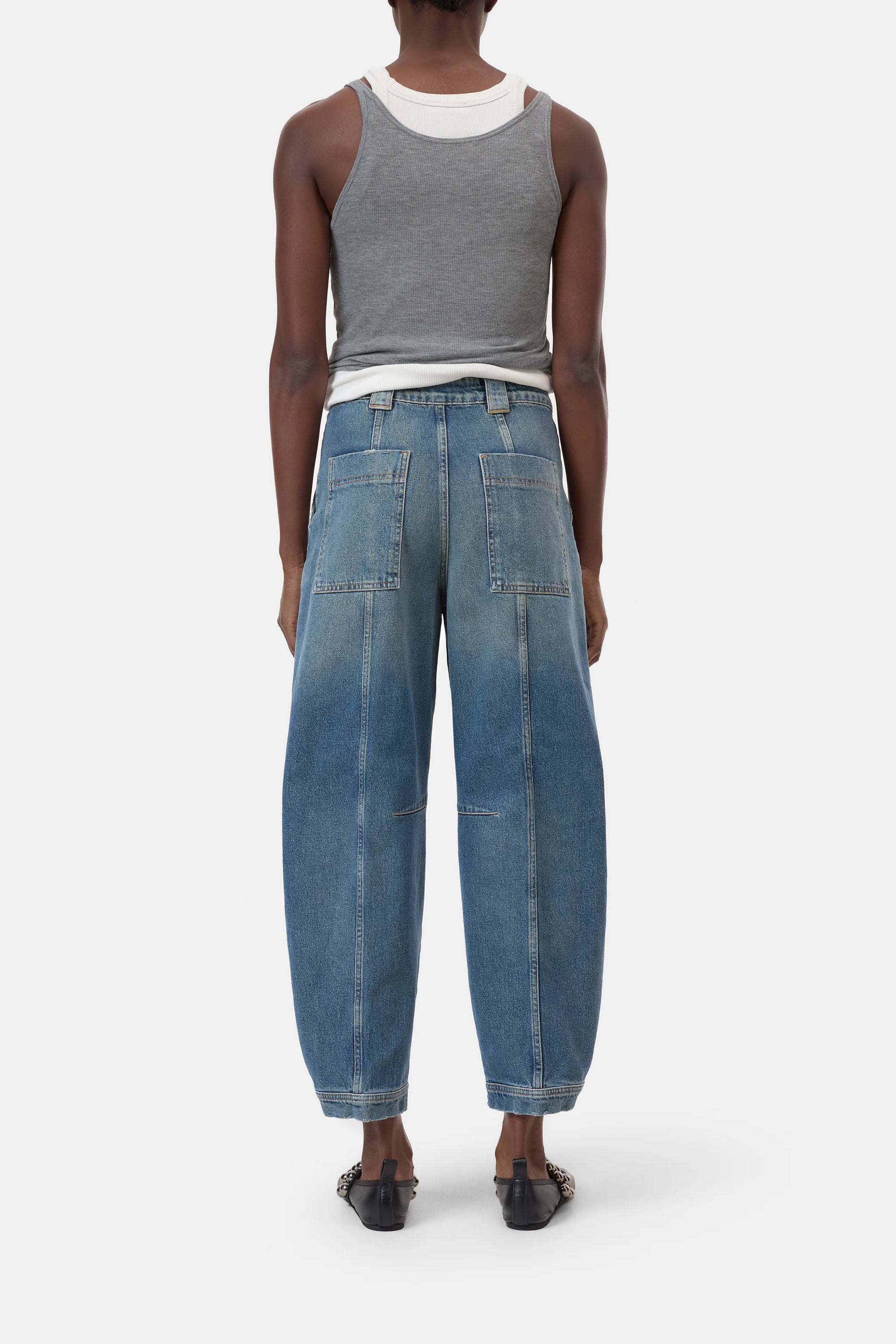A person faces away, wearing a gray tank over a white top, CLOSED Rhannon Denim vintage-wash barrel-fit jeans with large back pockets in comfort-stretch denim, and black slip-on shoes against a white background.