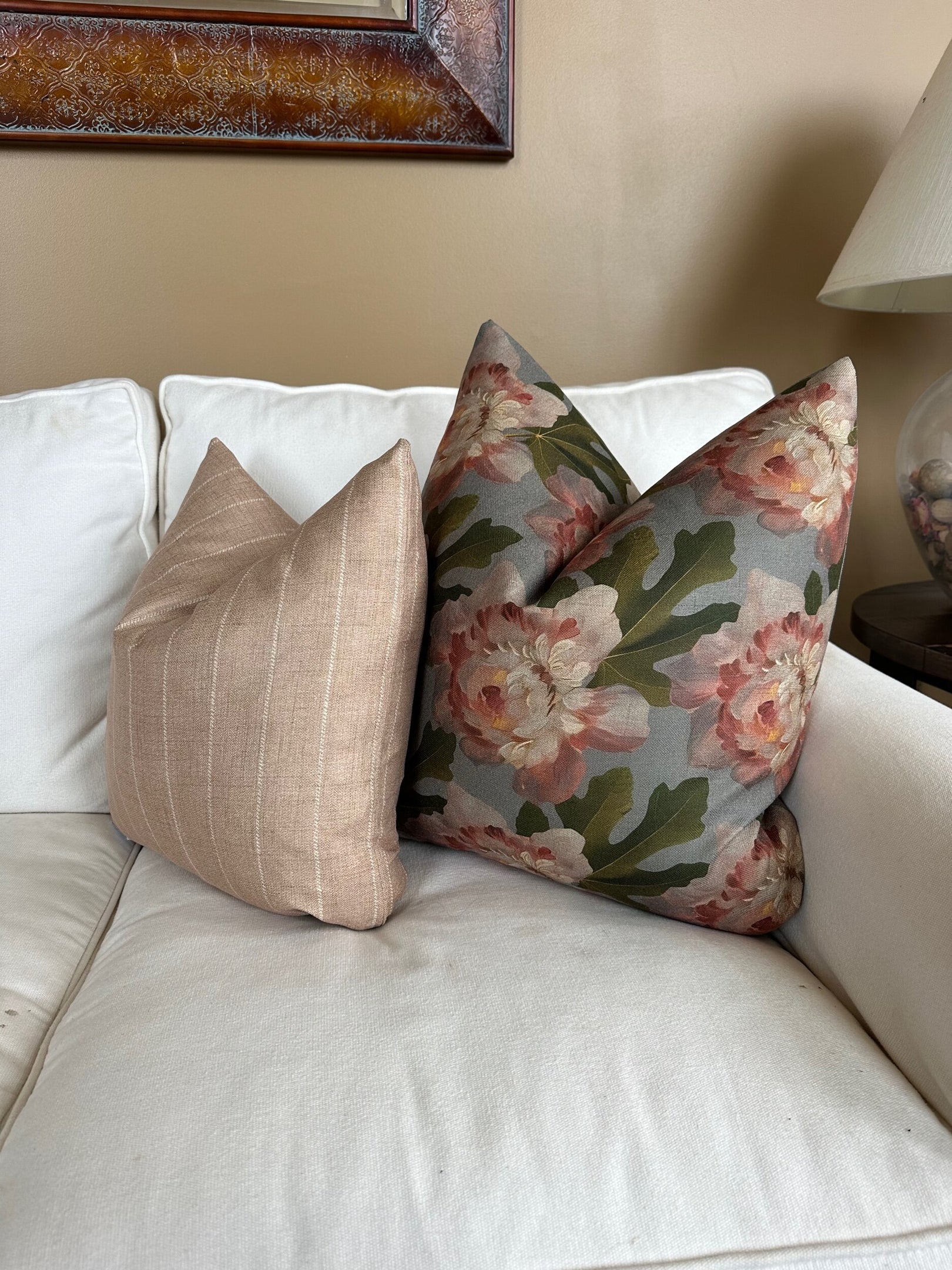 Two decorative Rennah Collective Iris Pillow 22x22 throw pillows rest on a white couch: one features light beige stripes in polyester-linen, the other displays pink flowers with green leaves. A lamp and framed art are seen in the background.