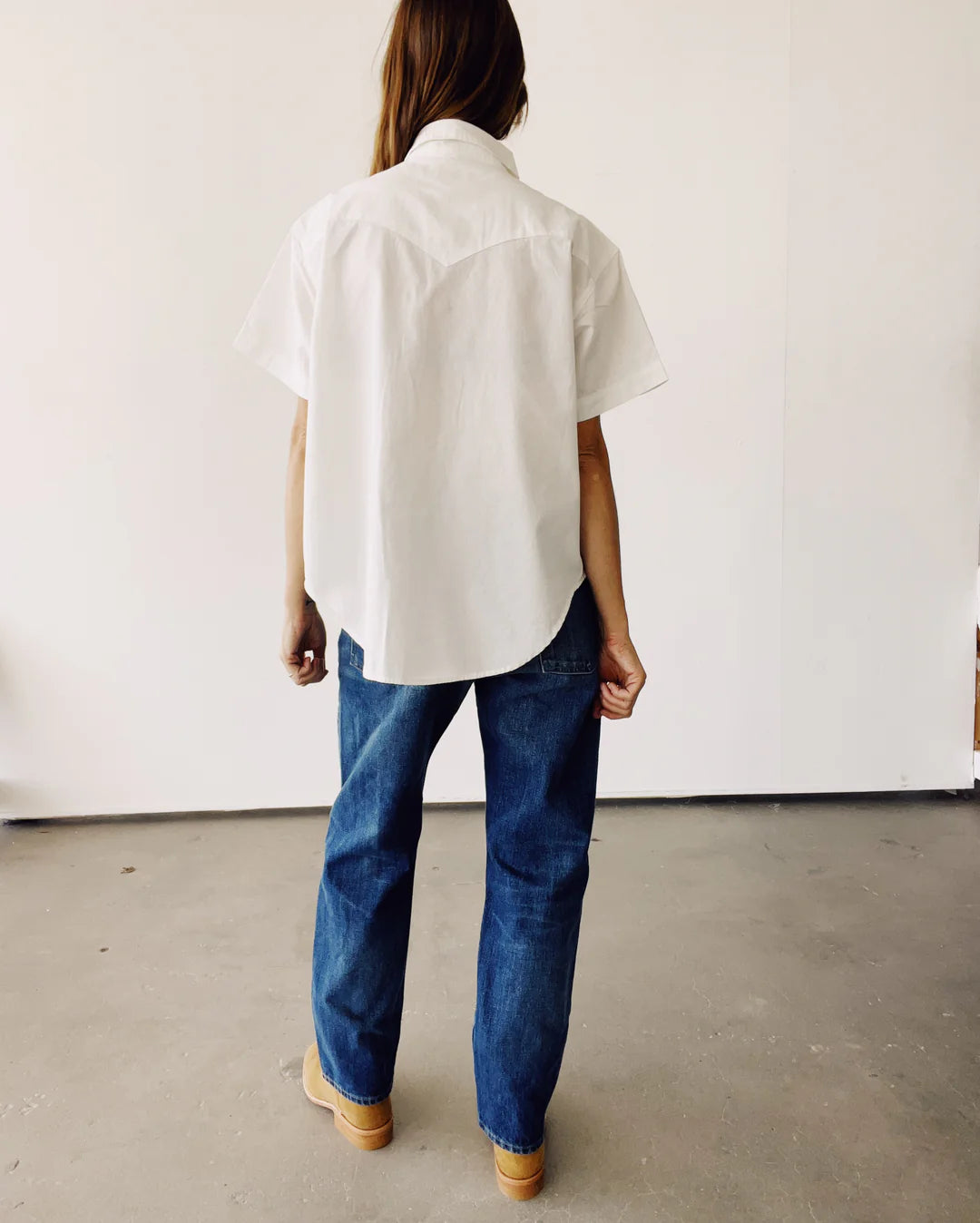 A person with long hair is facing away from the camera, dressed in a white oversized Western Shirt by Hey Gang, blue jeans, and tan shoes. They are positioned in a minimalistic indoor environment featuring a light-colored wall and gray floor.