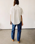 A person with long hair is facing away from the camera, dressed in a white oversized Western Shirt by Hey Gang, blue jeans, and tan shoes. They are positioned in a minimalistic indoor environment featuring a light-colored wall and gray floor.