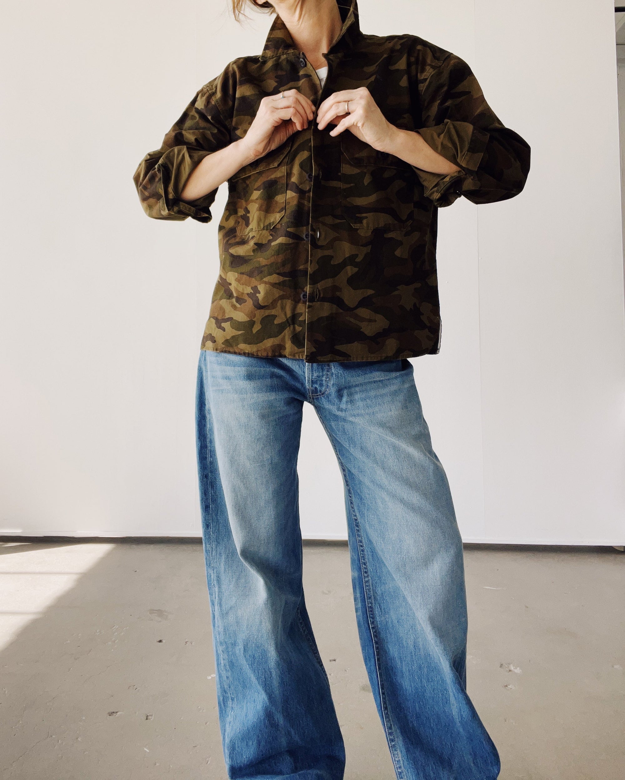 Someone stands indoors buttoning The Work Shirt by Hey Gang—a boxy fit camo jacket—paired with wide-leg blue jeans. Their face is hidden, and the setting features plain white walls and a gray floor.