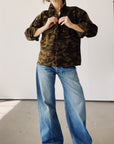 Someone stands indoors buttoning The Work Shirt by Hey Gang—a boxy fit camo jacket—paired with wide-leg blue jeans. Their face is hidden, and the setting features plain white walls and a gray floor.