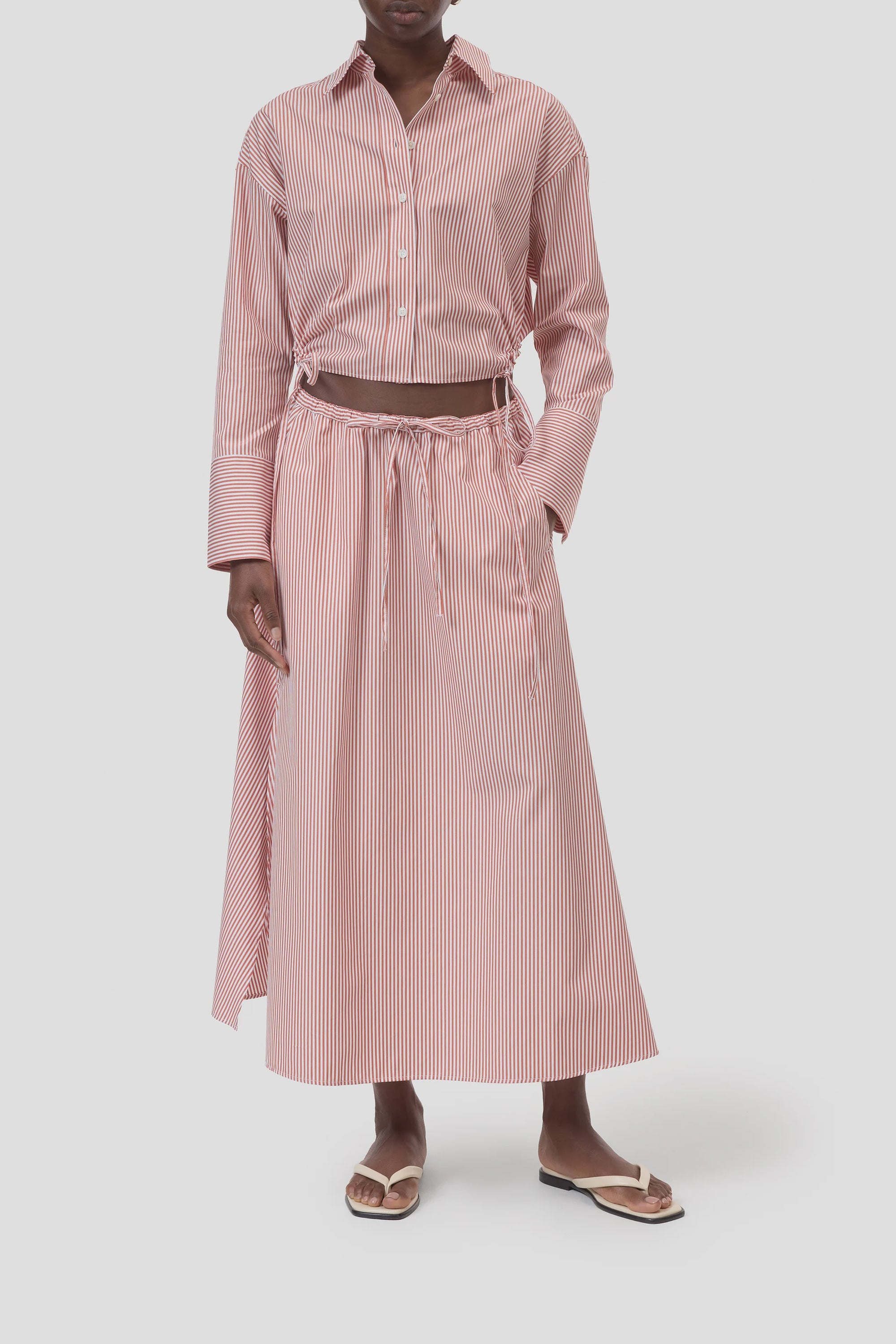 A person stands against a plain light background wearing a red and white vertically striped button-up shirt, the CLOSED Gathered Skirt with elasticated waistband (100% cotton), and white sandals.