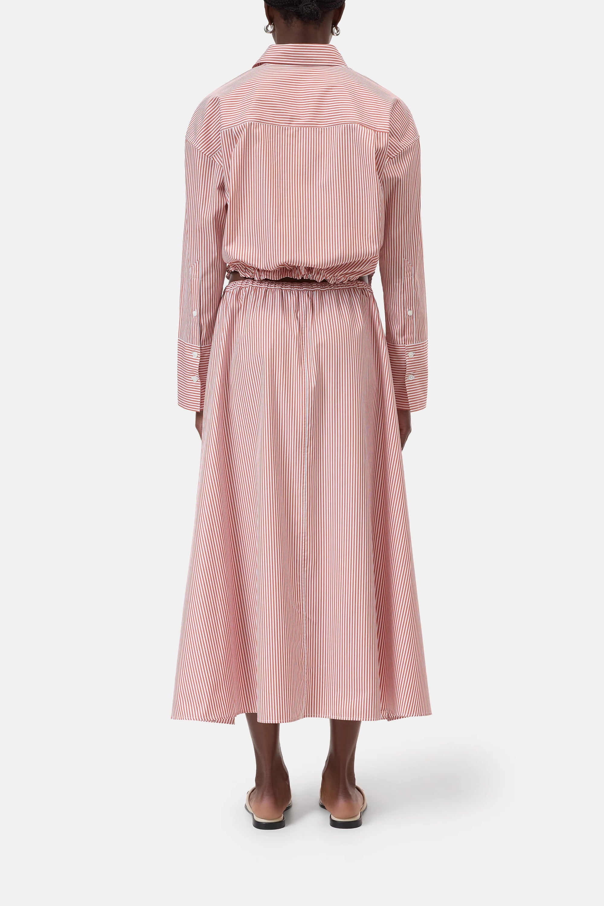 A person stands facing away, wearing CLOSED's Gathered Skirt—a long-sleeved, red-and-white vertically striped midi dress with a gathered skirt and elasticated waistband—paired with flat shoes against a plain white background.