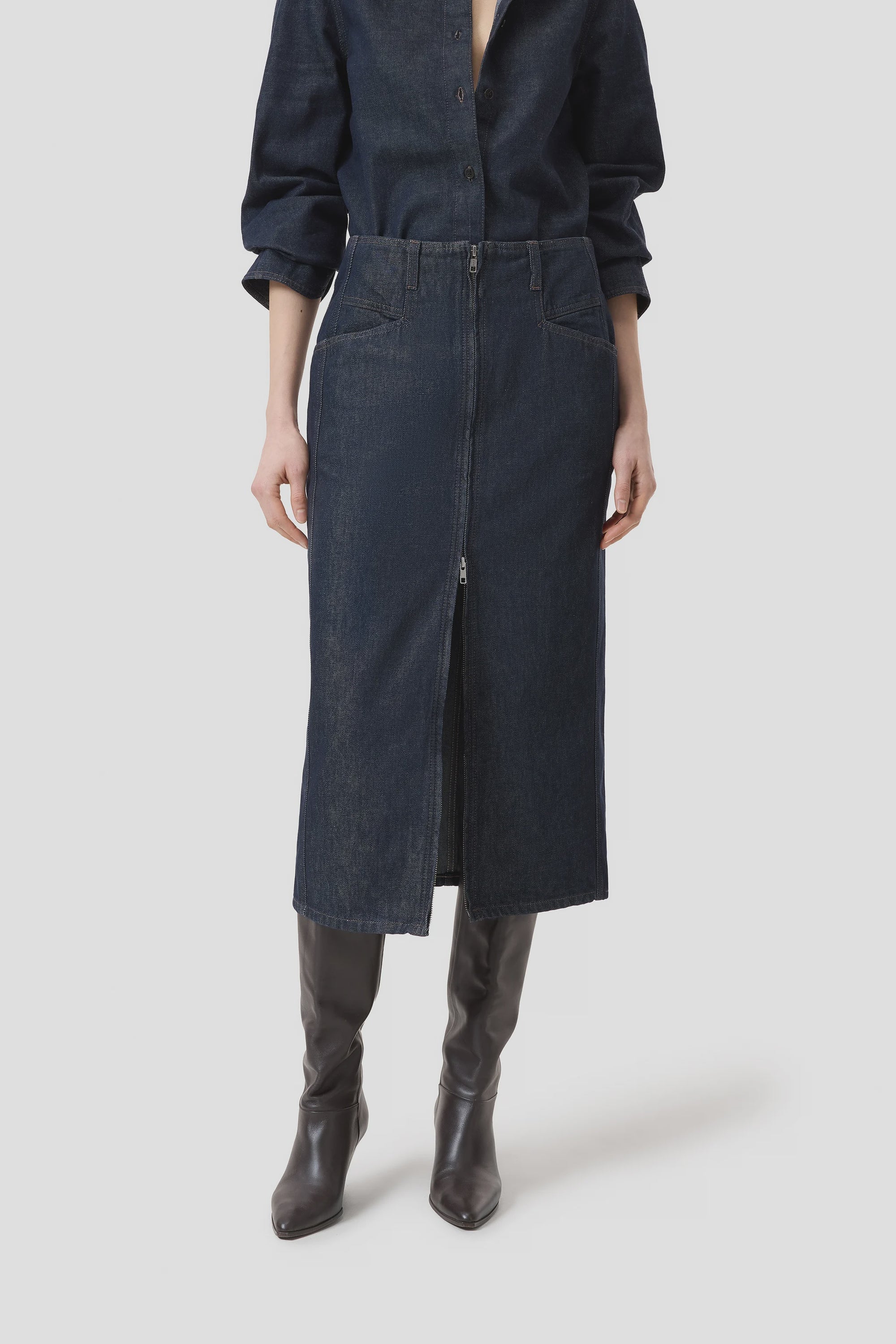 A person wears the CLOSED Zip Skirt in dark Italian Stay-Blue denim—a slim midi style—paired with a matching denim shirt and knee-high brown leather boots, standing against a plain light background.