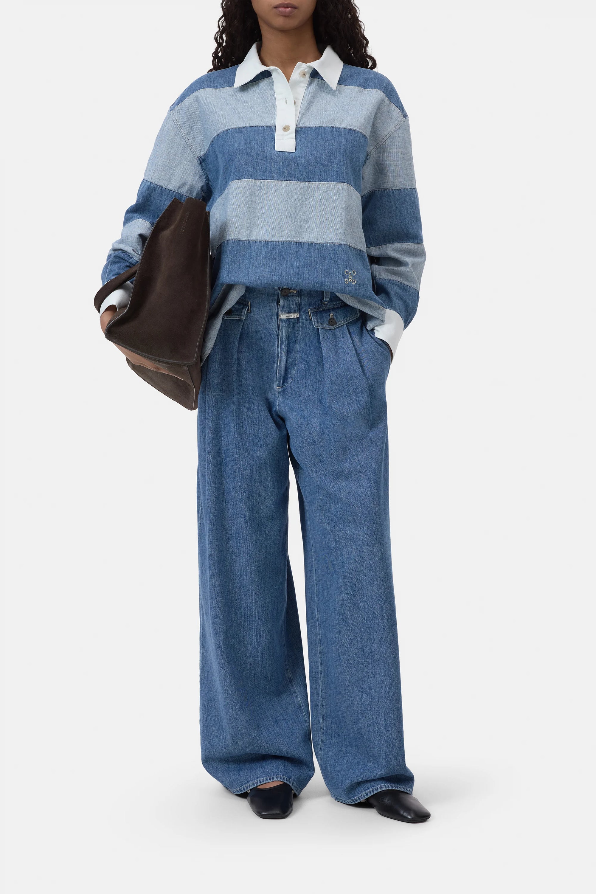 Wearing a CLOSED Rugby Shirt in blue and light blue stripes tucked into wide-leg lightweight jeans, this person holds a large dark brown bag, has hands in pockets, and wears black shoes.