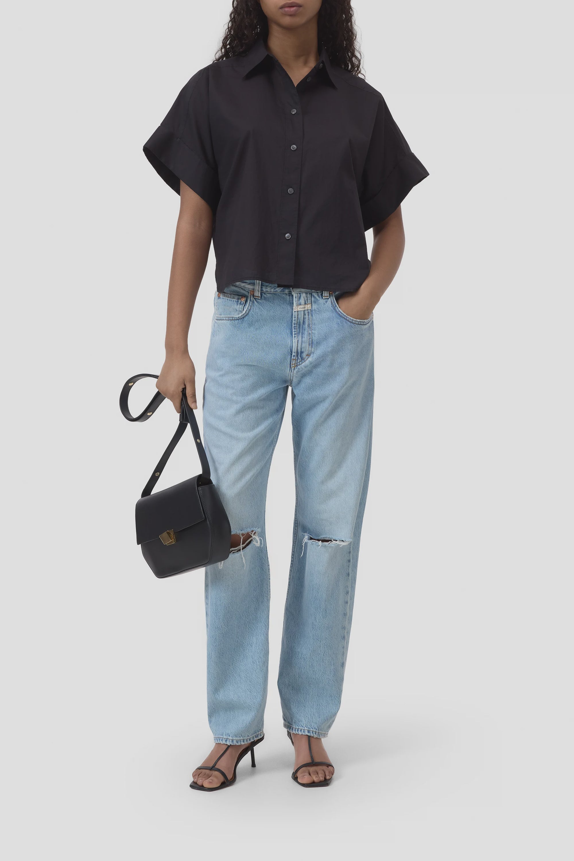 A woman wears the CLOSED Turn-Up Shirt, a relaxed fit cotton style with mother-of-pearl buttons, paired with light blue ripped jeans, black heeled sandals, and a small black handbag. Her left hand is in her pocket; her face is partly out of frame.