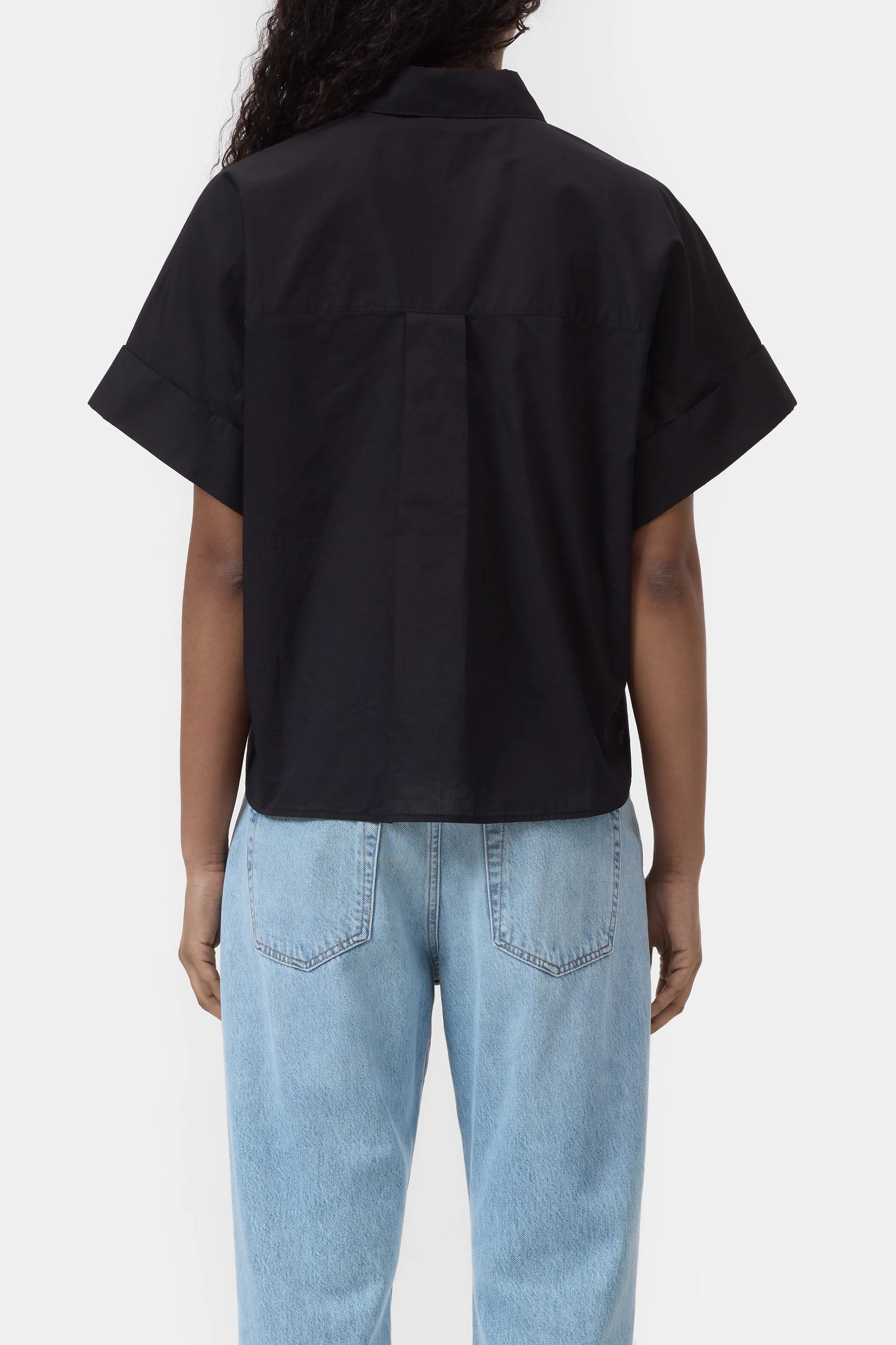 A person with long, curly hair is shown from the back, wearing the CLOSED Turn-Up Shirt in black with a relaxed fit and short sleeves, paired with light blue high-waisted jeans against a plain white background.
