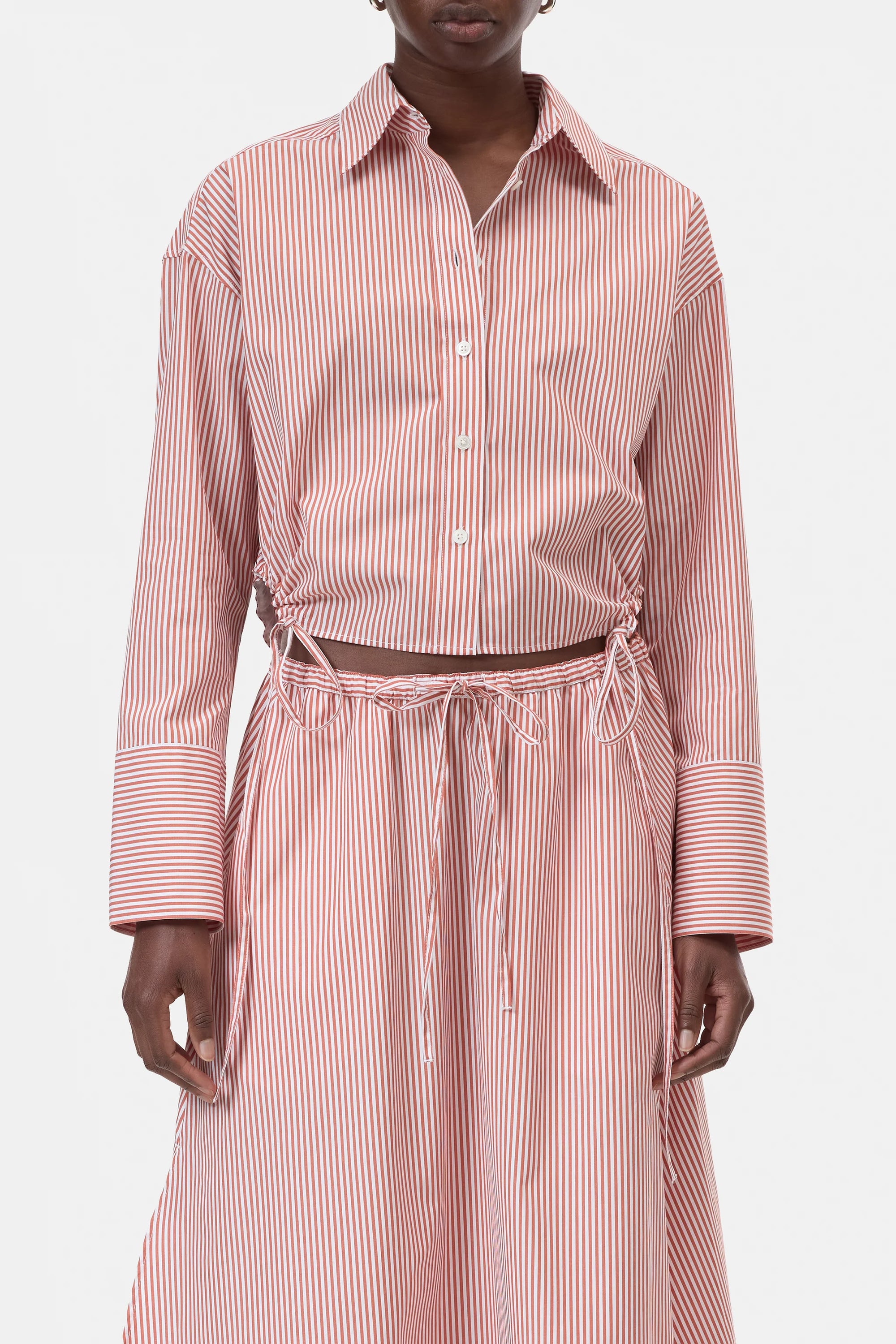 A person wears the CLOSED Gathered Shirt, a relaxed-fit women's blouse in pink and white vertical stripes with wide cuffs, paired with a matching high-waisted drawstring skirt. The coordinated set has a laid-back vibe, showing only the torso and upper legs.