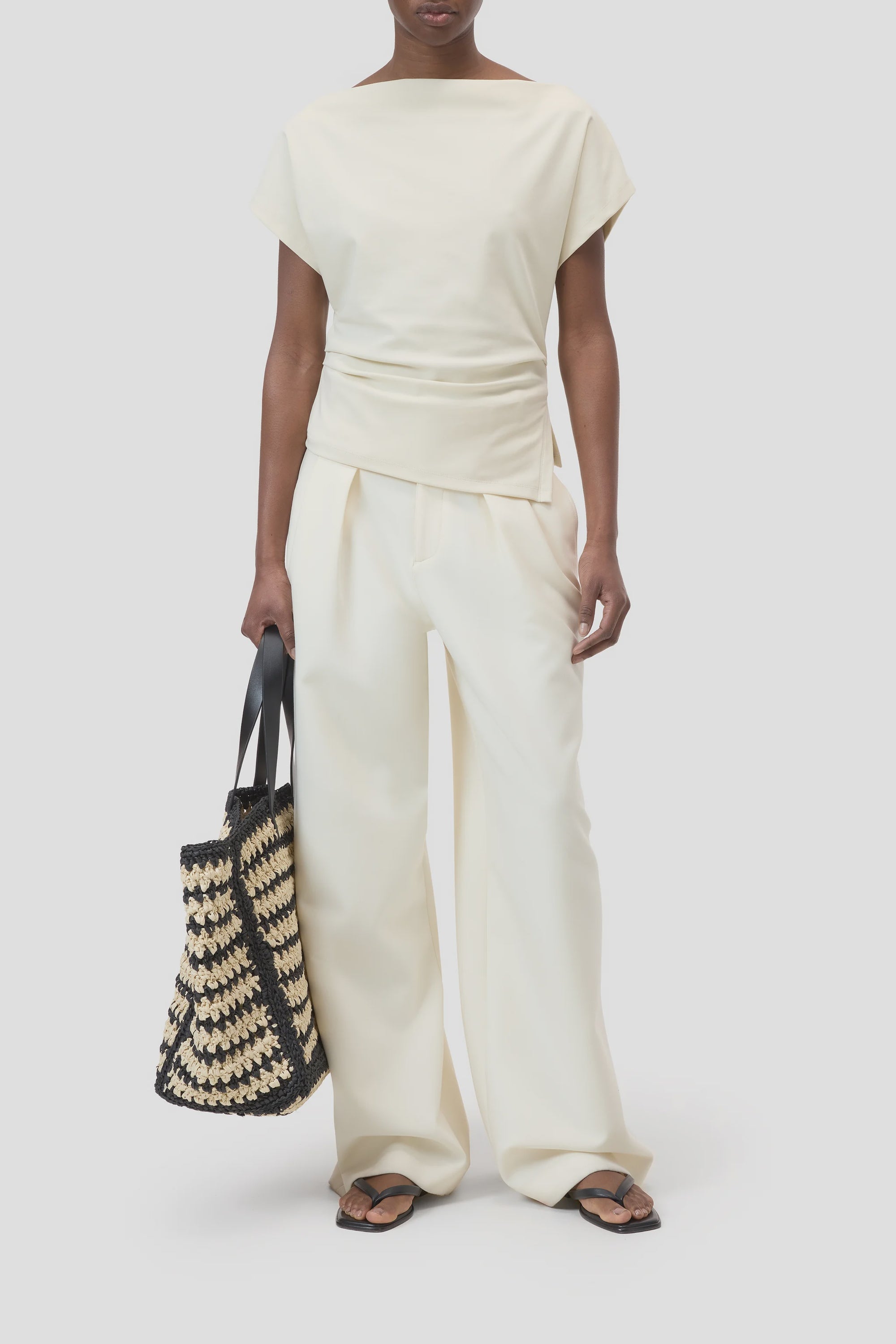 A person wears the CLOSED Off Shoulder Top in cream slim-fit cotton-elastane with wide-leg pants, holding a woven black and beige tote bag and wearing black flip-flops against a plain light gray background.