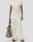 A person wears the CLOSED Off Shoulder Top in cream slim-fit cotton-elastane with wide-leg pants, holding a woven black and beige tote bag and wearing black flip-flops against a plain light gray background.