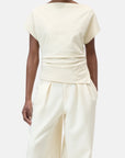 A person wears the CLOSED Off Shoulder Top in slim fit off-white cotton-elastane with a draped waist, paired with matching high-waisted wide-leg trousers, standing against a plain light background.