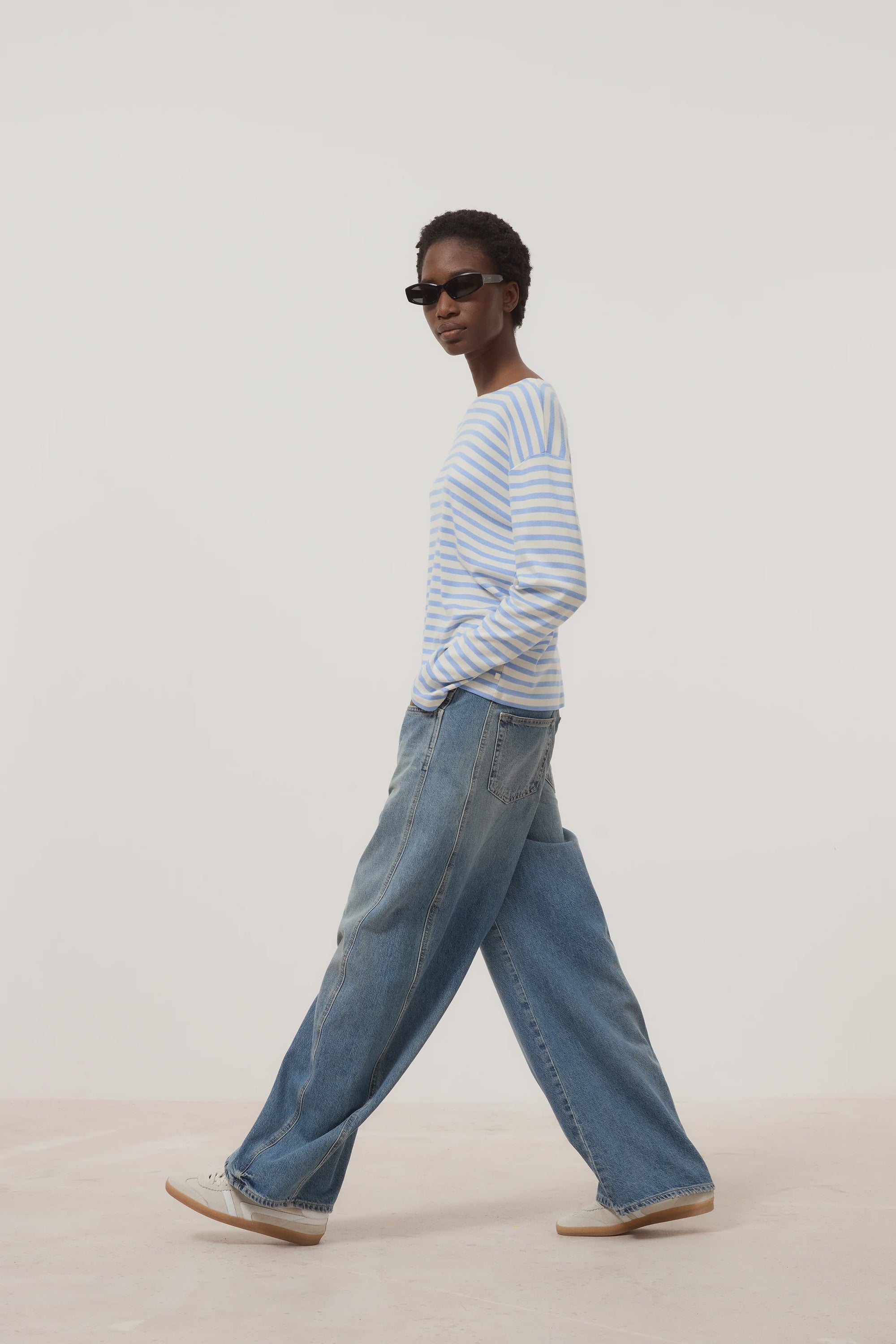 A person in dark sunglasses, wide-leg blue jeans, white sneakers, and the CLOSED Basic Long Sleeve Tee with light blue and white stripes walks against a plain light background.