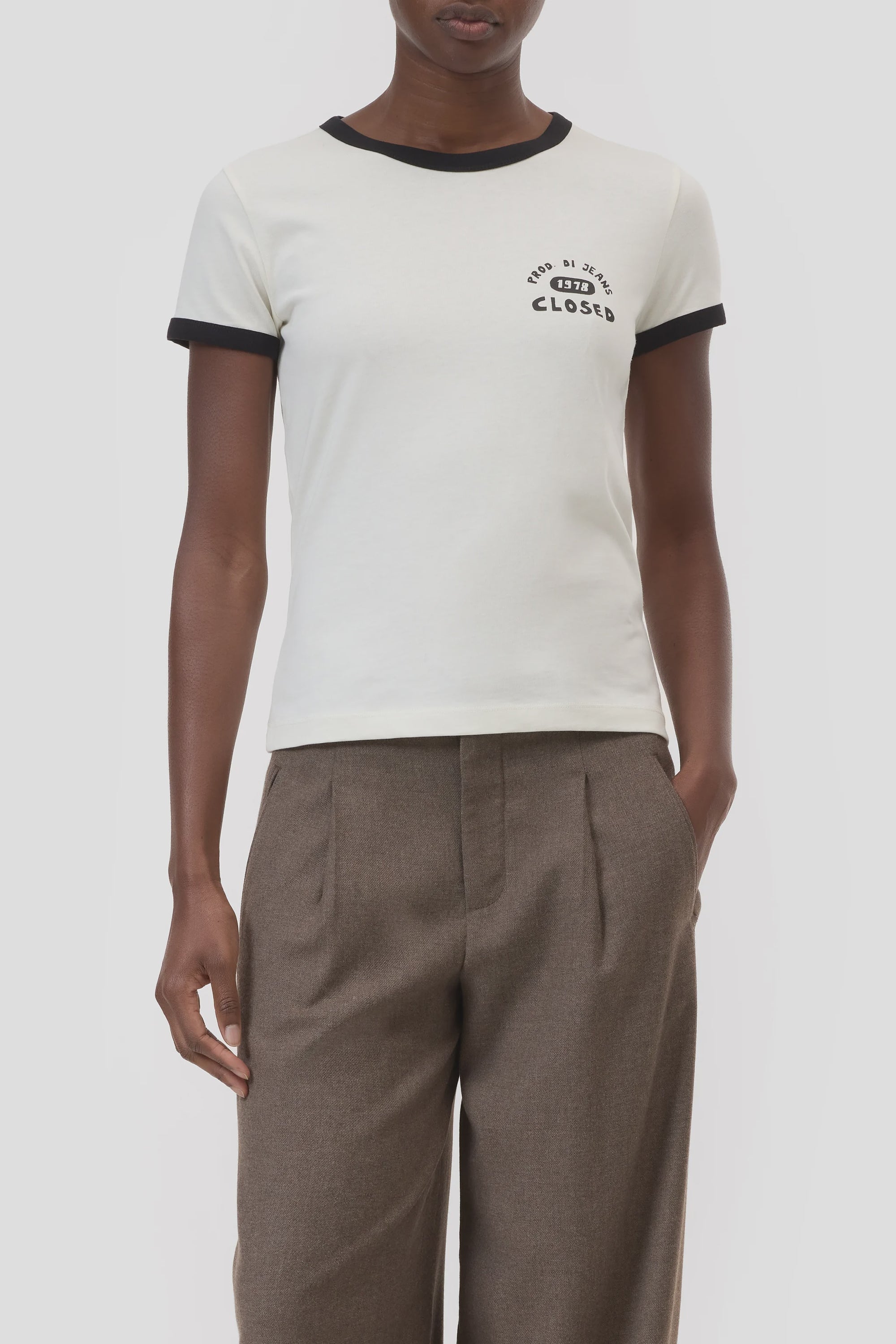 A person wears the CLOSED Slim T-Shirt—white with black collar and sleeve cuffs, featuring a small "Closed" logo on the chest—paired with brown pleated trousers against a plain background.