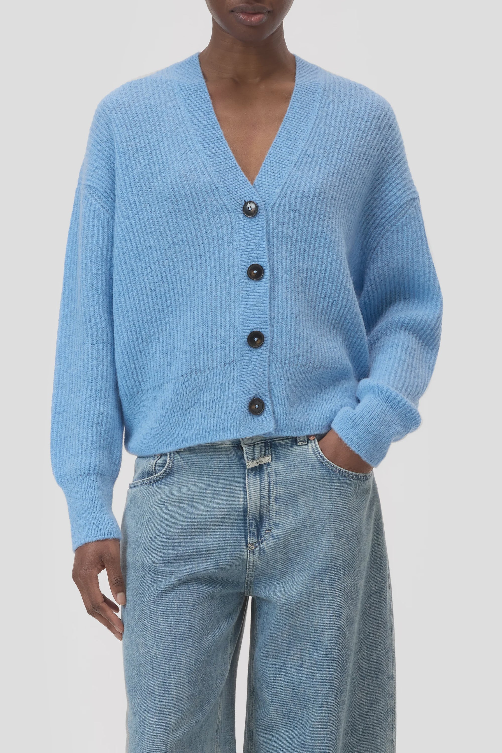 A person wearing a light blue CLOSED V Neck Cardigan with long sleeves and ribbed texture, paired with light blue high-waisted jeans. Their left hand is in their pocket.
