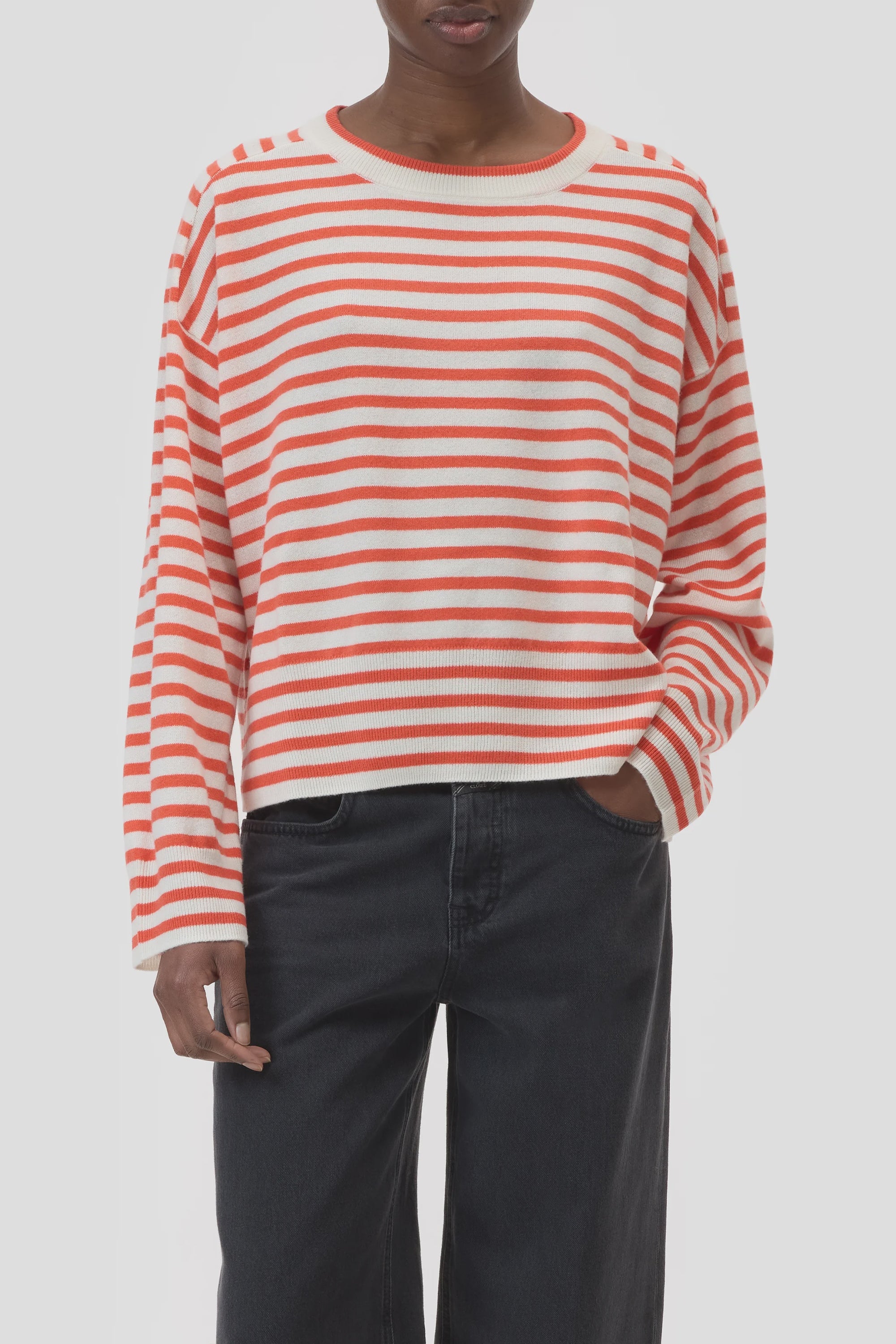 A person wearing the CLOSED Striped Crew Neck, a loose-fitting orange and white sweater, with dark high-waisted jeans stands with one hand in their pocket against a plain background.