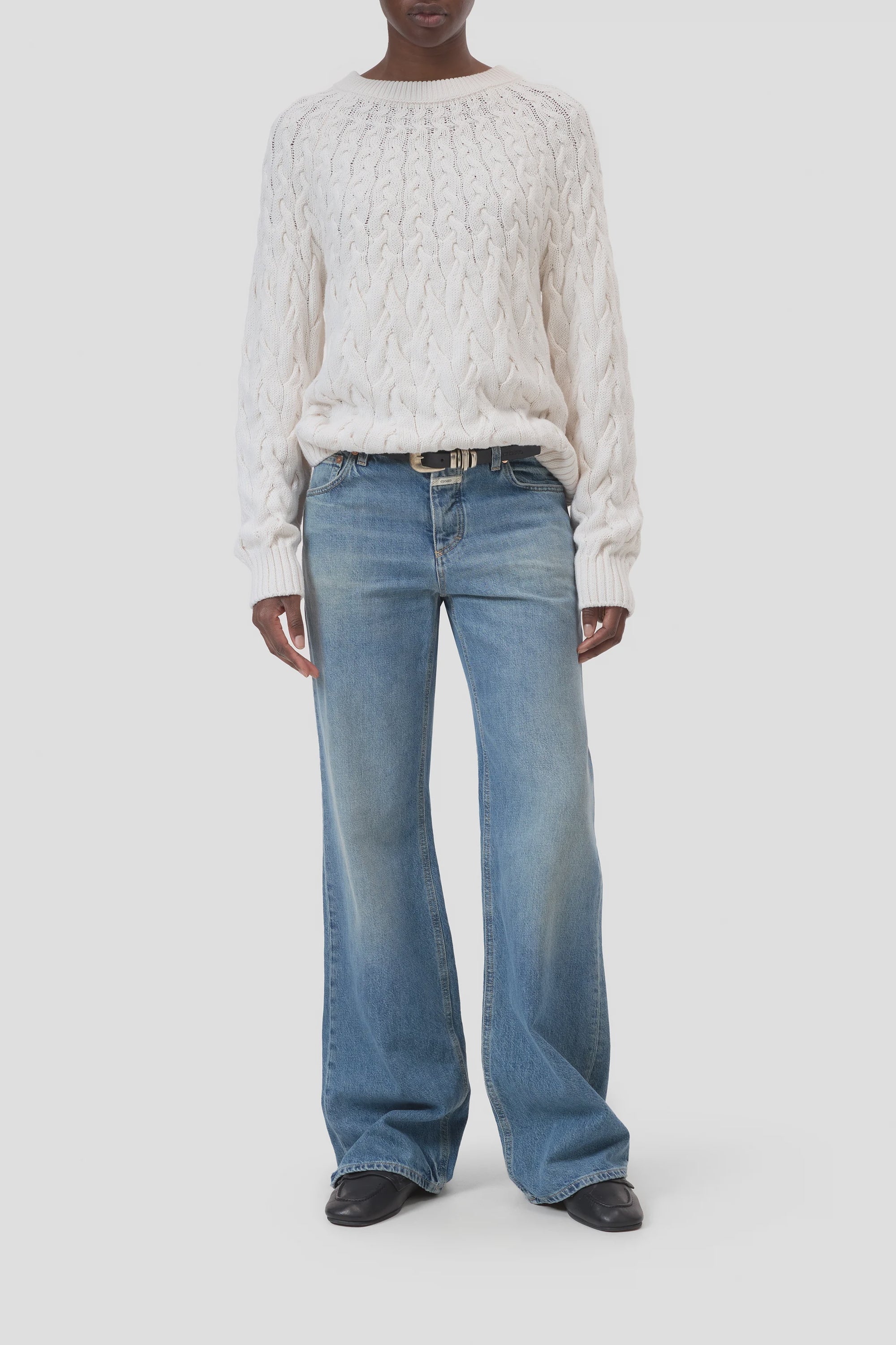 A person wears the CLOSED Crew Neck Cable Knit sweater, blue wide-leg jeans, a black belt, and black shoes while standing against a plain white background.