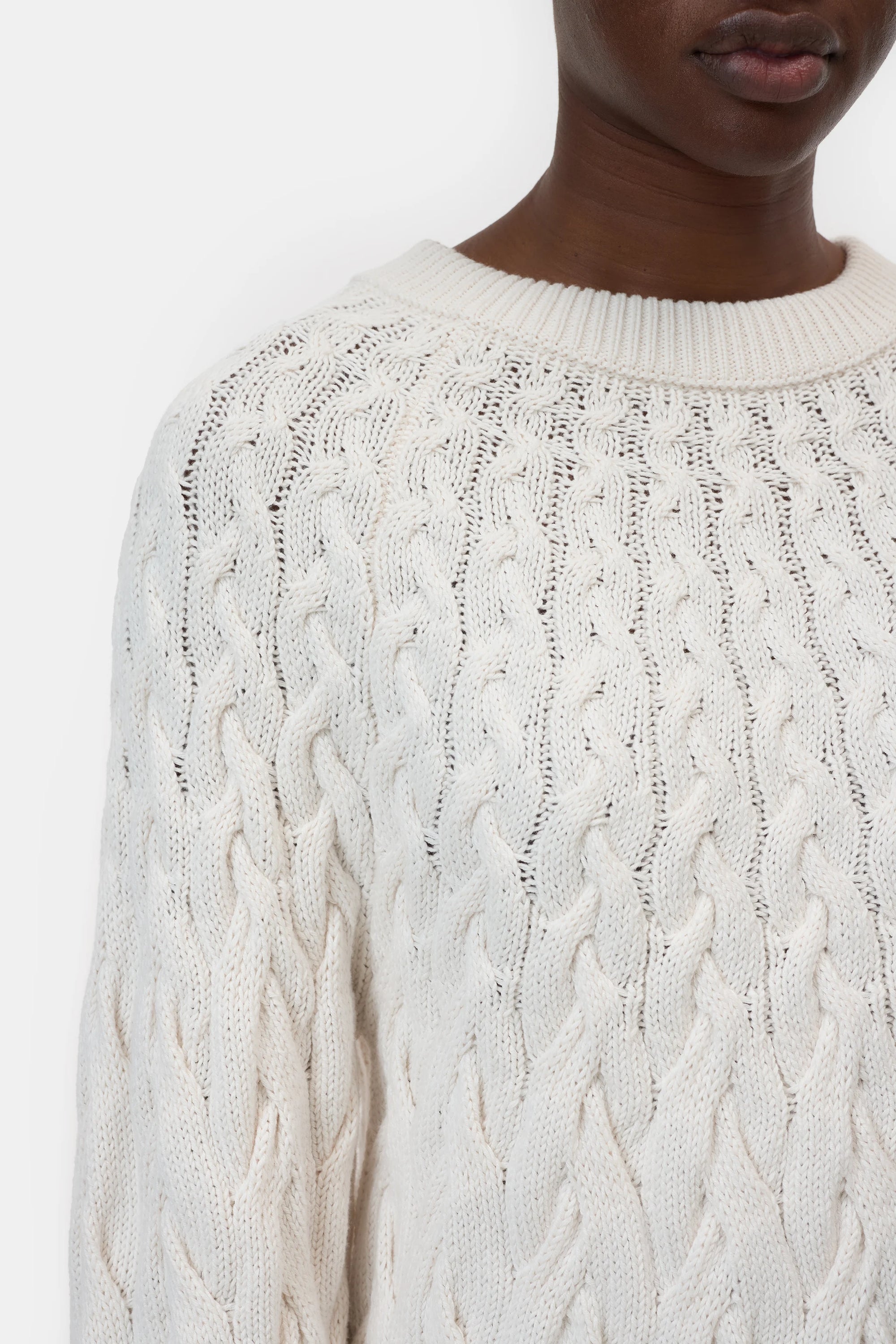 Close-up of a person wearing the CLOSED Crew Neck Cable Knit sweater in off-white, showing their lower face, neck, and upper torso against a plain white background. Ribbed neckline and chunky cable knit details are visible.