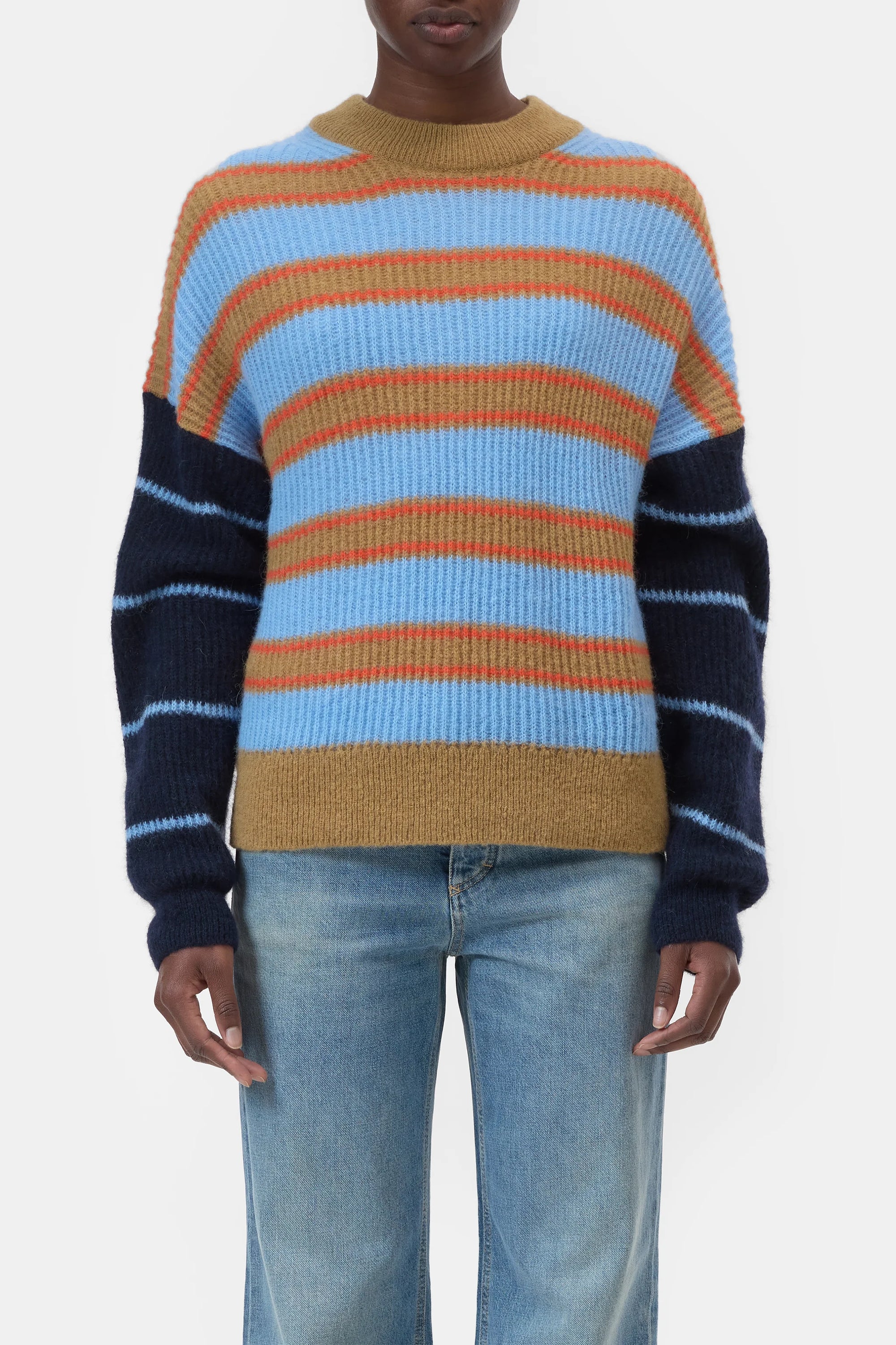 A person models the CLOSED Crewneck LS Knit, a colorful striped wool sweater in blue, red, mustard, and navy, paired with light blue jeans against a plain white background.