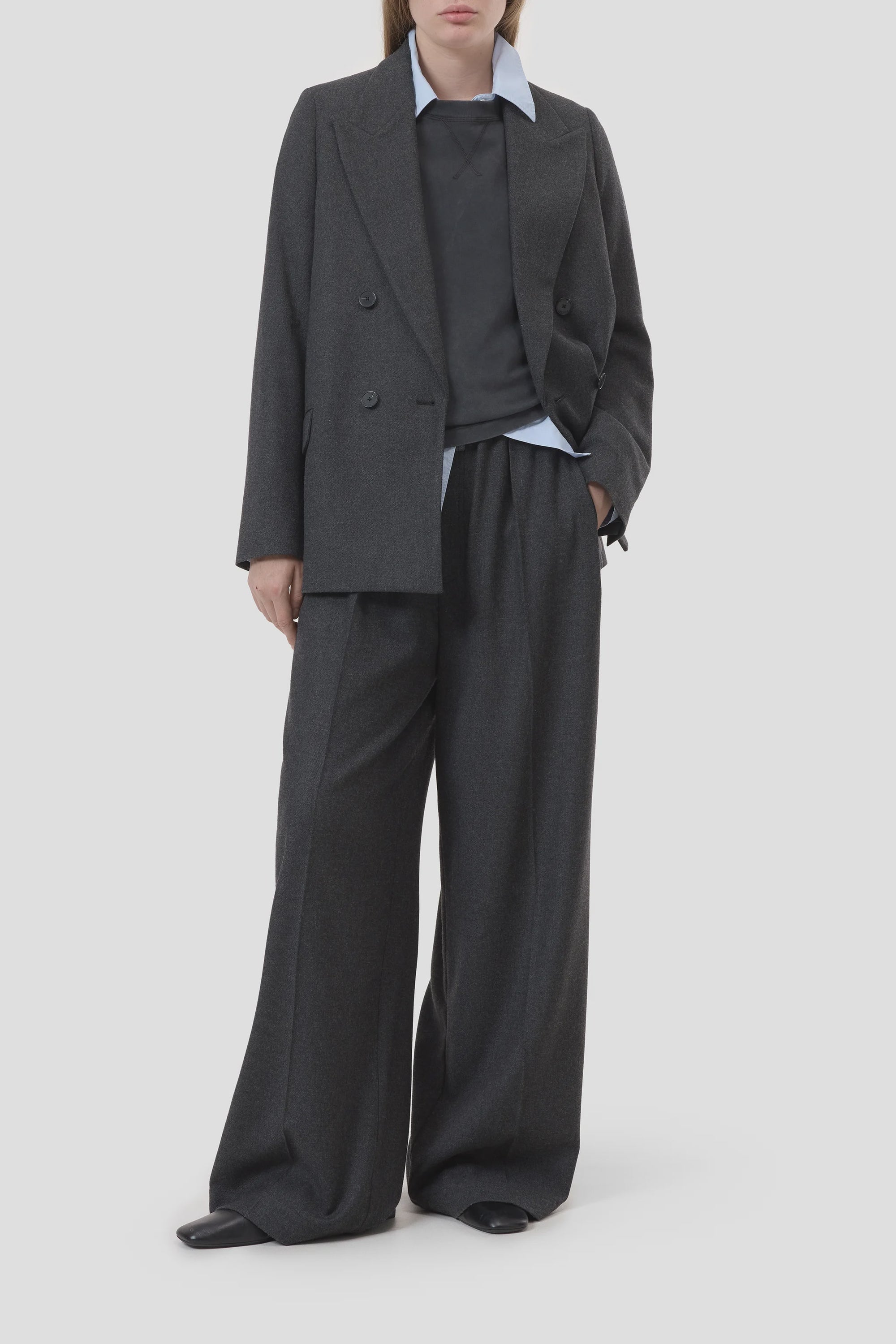 A person wears CLOSED’s Fitted Doublebreasted Blazer in dark gray with wide-leg trousers over a light blue collared shirt, standing against a plain light background. Their face is partially out of frame.