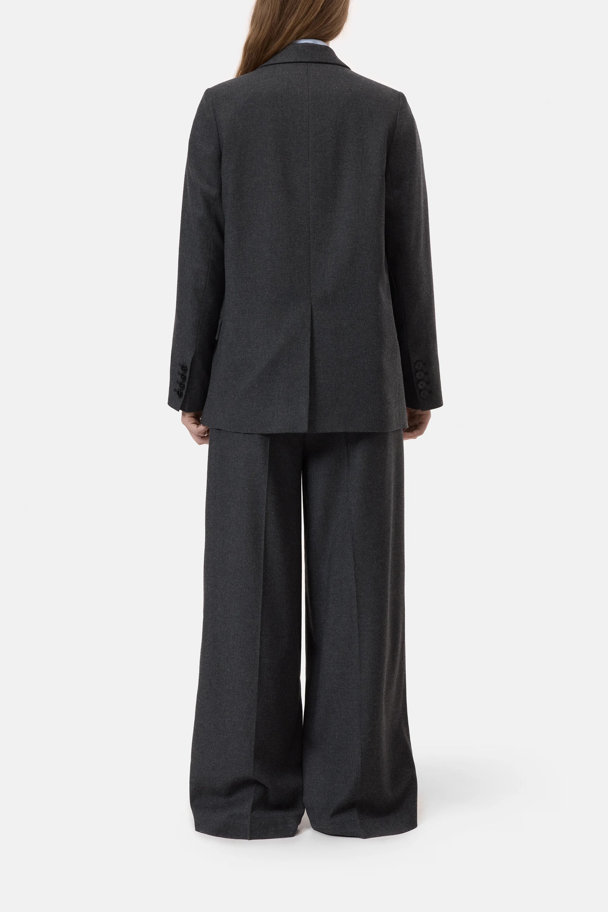 A person with long brown hair stands facing away against a plain white background, wearing CLOSED’s Fitted Doublebreasted Blazer in dark gray paired with wide-leg trousers.