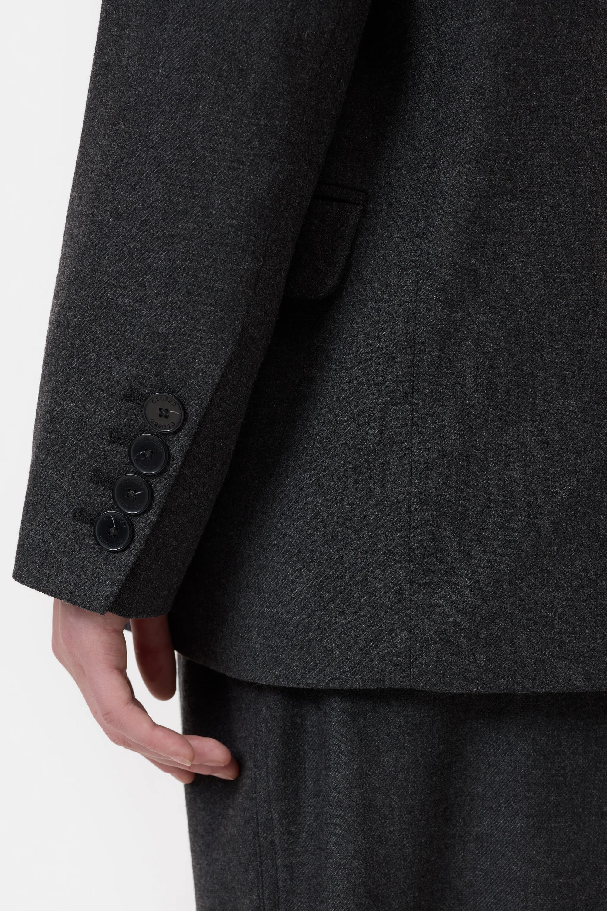 A close-up shows a person wearing the CLOSED Fitted Doublebreasted Blazer in dark gray, highlighting the sleeve with four black buttons and part of their hand against a plain, light background.