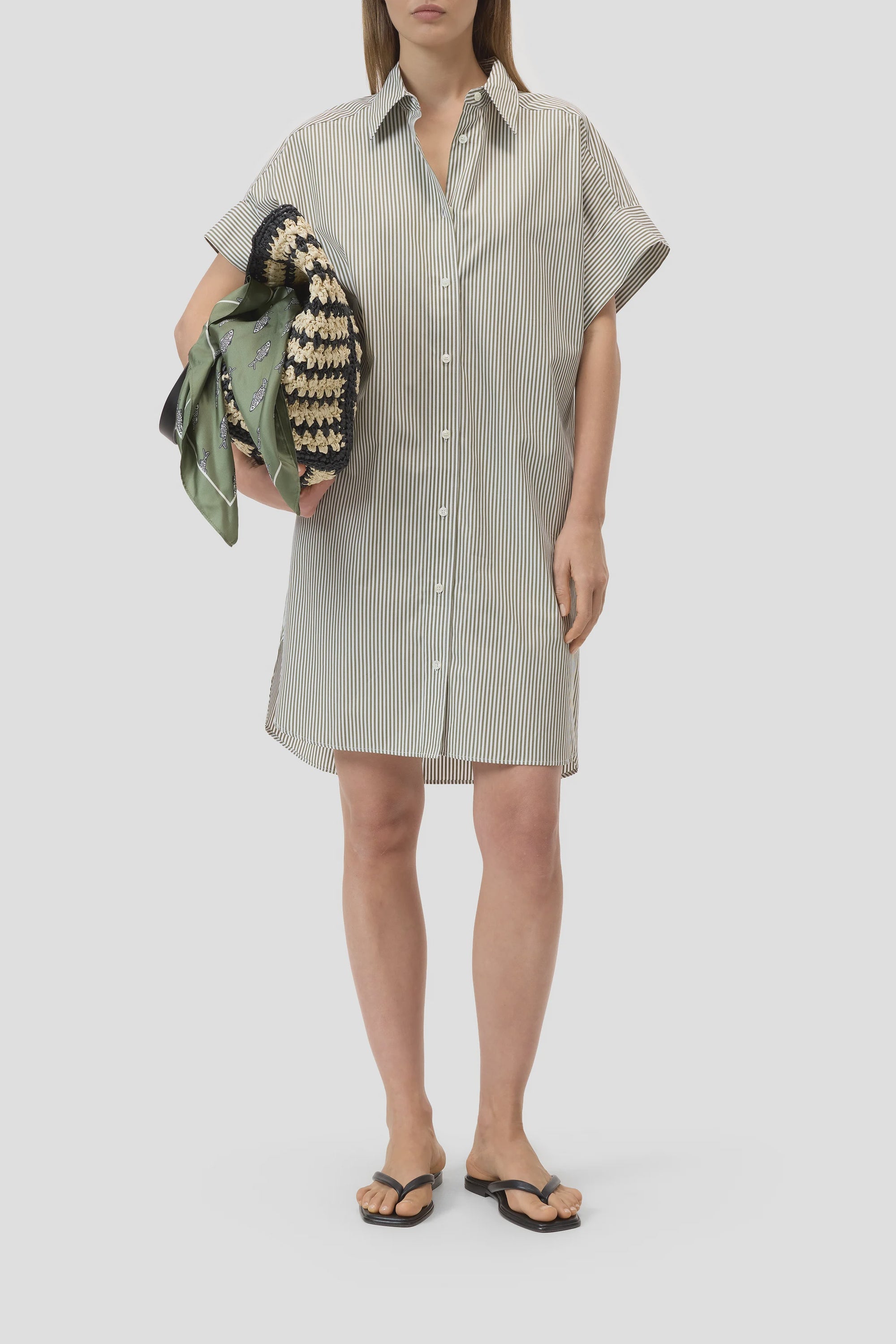 A person in the CLOSED Turn-up Shirtdress with short sleeves, stripes, and mother-of-pearl buttons, paired with flip-flops and holding a green and beige woven bag draped with scarves, stands against a plain light background.