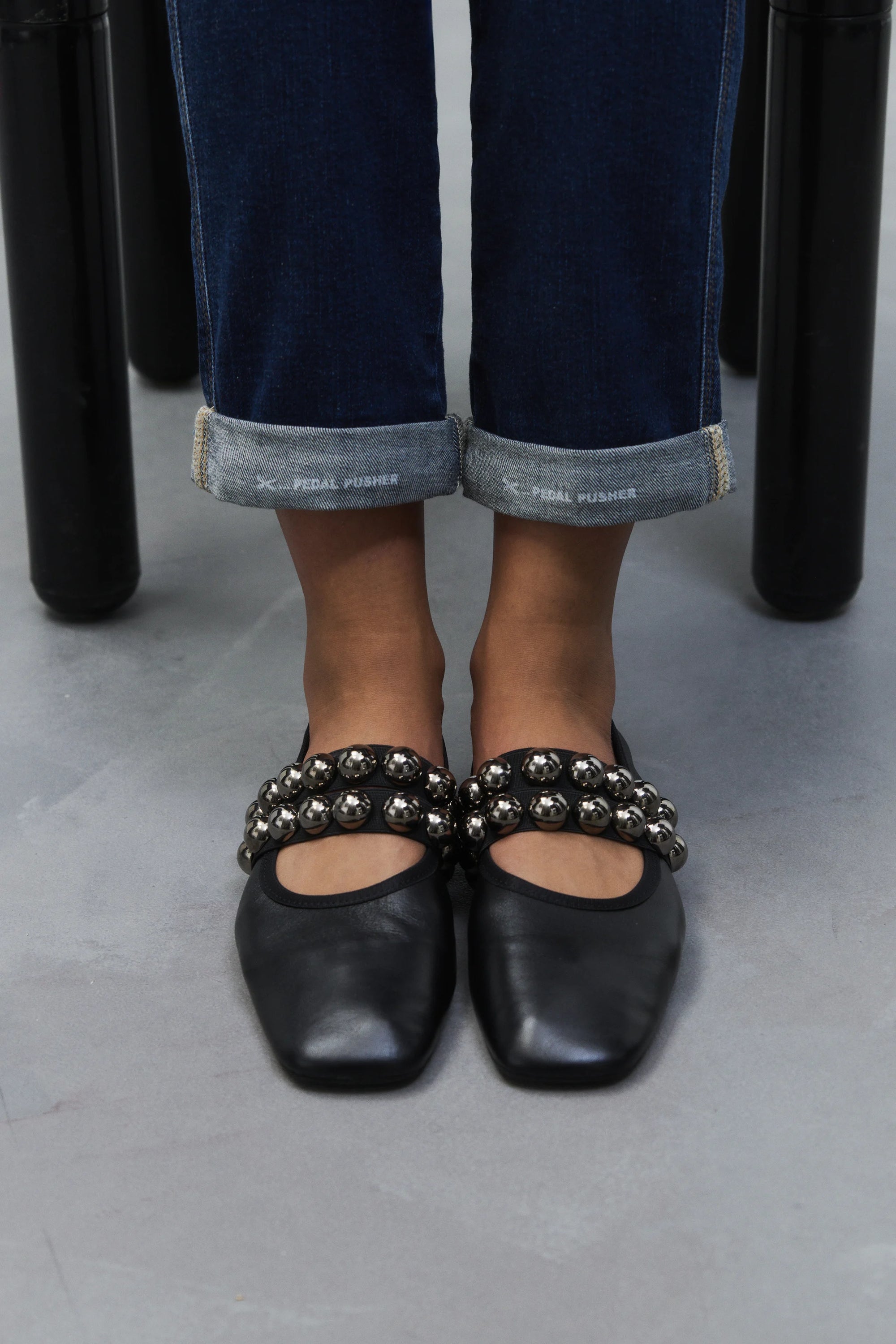 A person in cuffed blue jeans wears CLOSED Ballerina black square-toe flats with silver studs, leather insoles, and made in Italy, while standing on a gray floor.