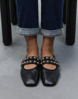 A person in cuffed blue jeans wears CLOSED Ballerina black square-toe flats with silver studs, leather insoles, and made in Italy, while standing on a gray floor.