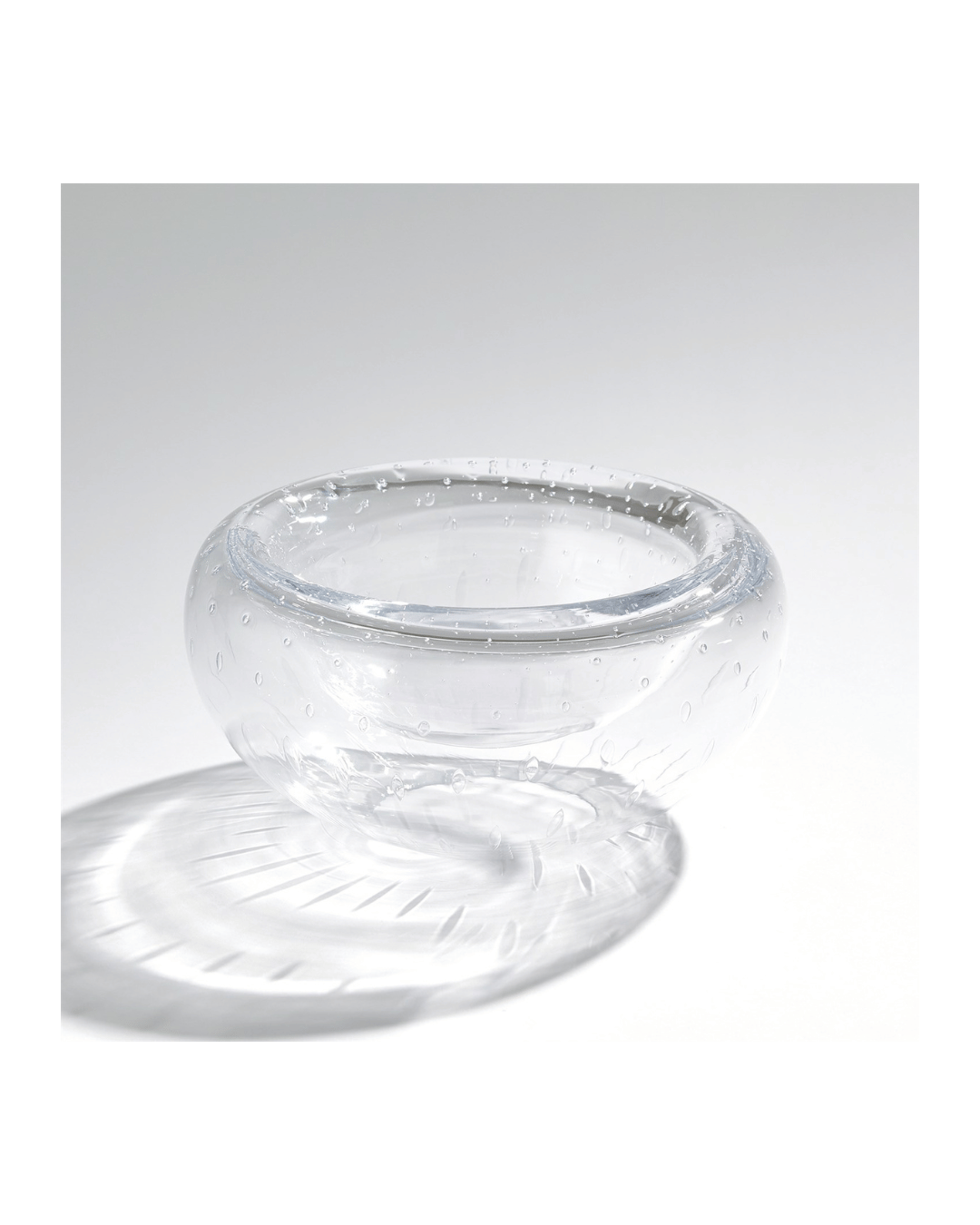The Double Take Bowl-Clear Seeded-Medium by Global Views, a clear spherical glass bowl with a subtle dimpled texture, casts a soft shadow on a white surface. Its finish beautifully complements the plain, evenly lit background.
