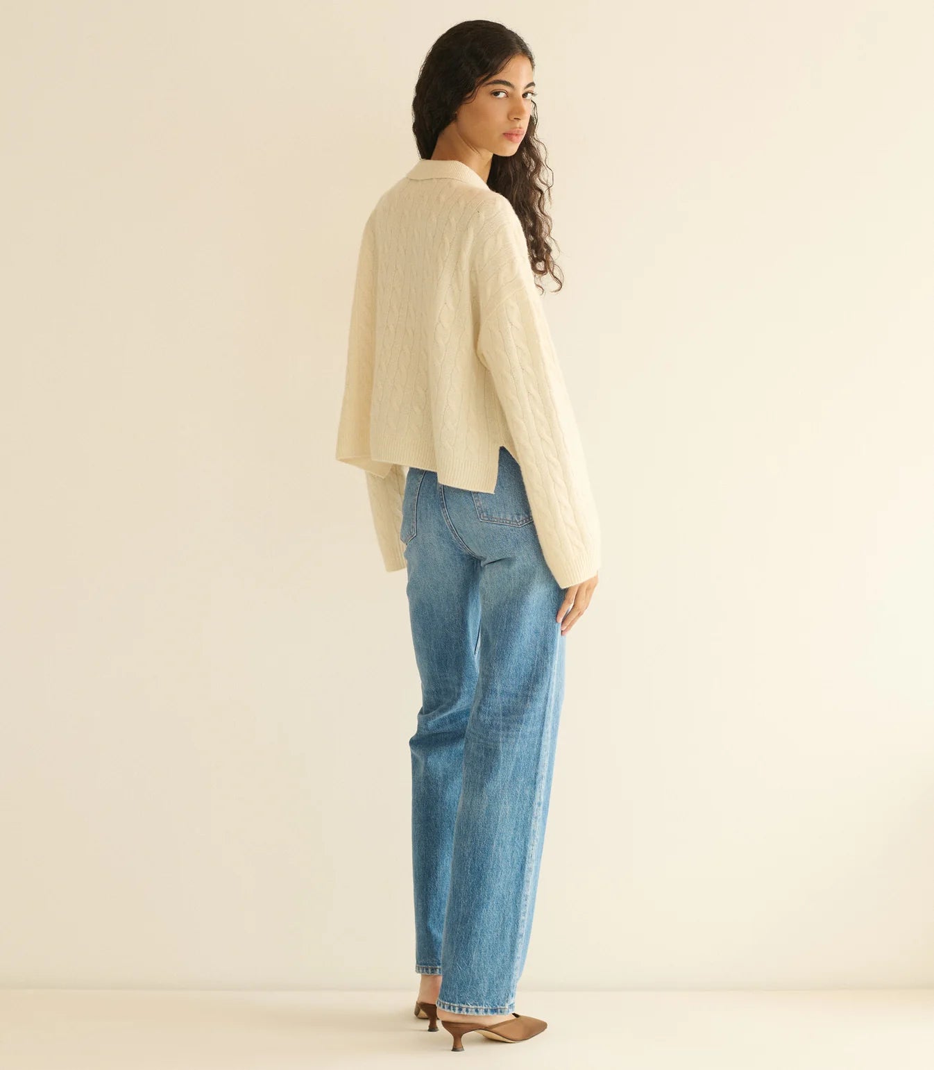 A person with long curly hair, wearing the DOEN Cambridge Cashmere Sweater in cream, blue jeans, and brown heels stands sideways and looks over their shoulder against a plain light background.