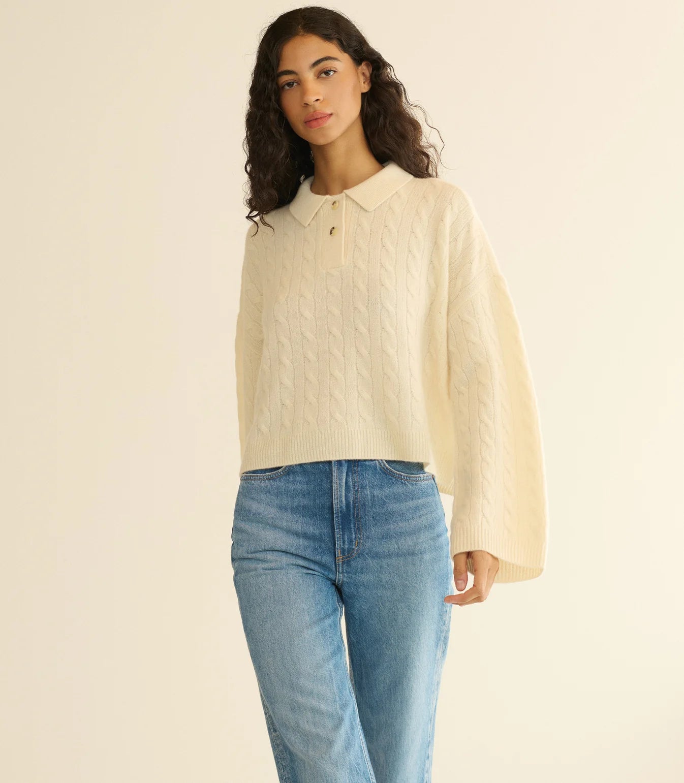 A woman with long curly hair wears the DOEN Cambridge Cashmere Sweater in cream, featuring a cable knit pattern and Henley collar, paired with blue jeans. She stands against a plain, light background.