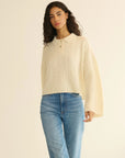 A woman with long curly hair wears the DOEN Cambridge Cashmere Sweater in cream, featuring a cable knit pattern and Henley collar, paired with blue jeans. She stands against a plain, light background.