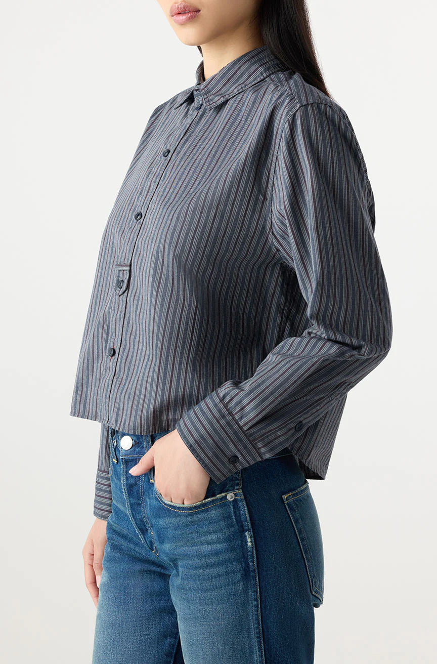 A woman wears the Lena Shirt by AMO Denim, a gray and blue striped, long-sleeve button-up with a relaxed fit, paired with blue jeans. She stands sideways, one hand in her pocket, against a plain white background.