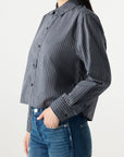 A woman wears the Lena Shirt by AMO Denim, a gray and blue striped, long-sleeve button-up with a relaxed fit, paired with blue jeans. She stands sideways, one hand in her pocket, against a plain white background.