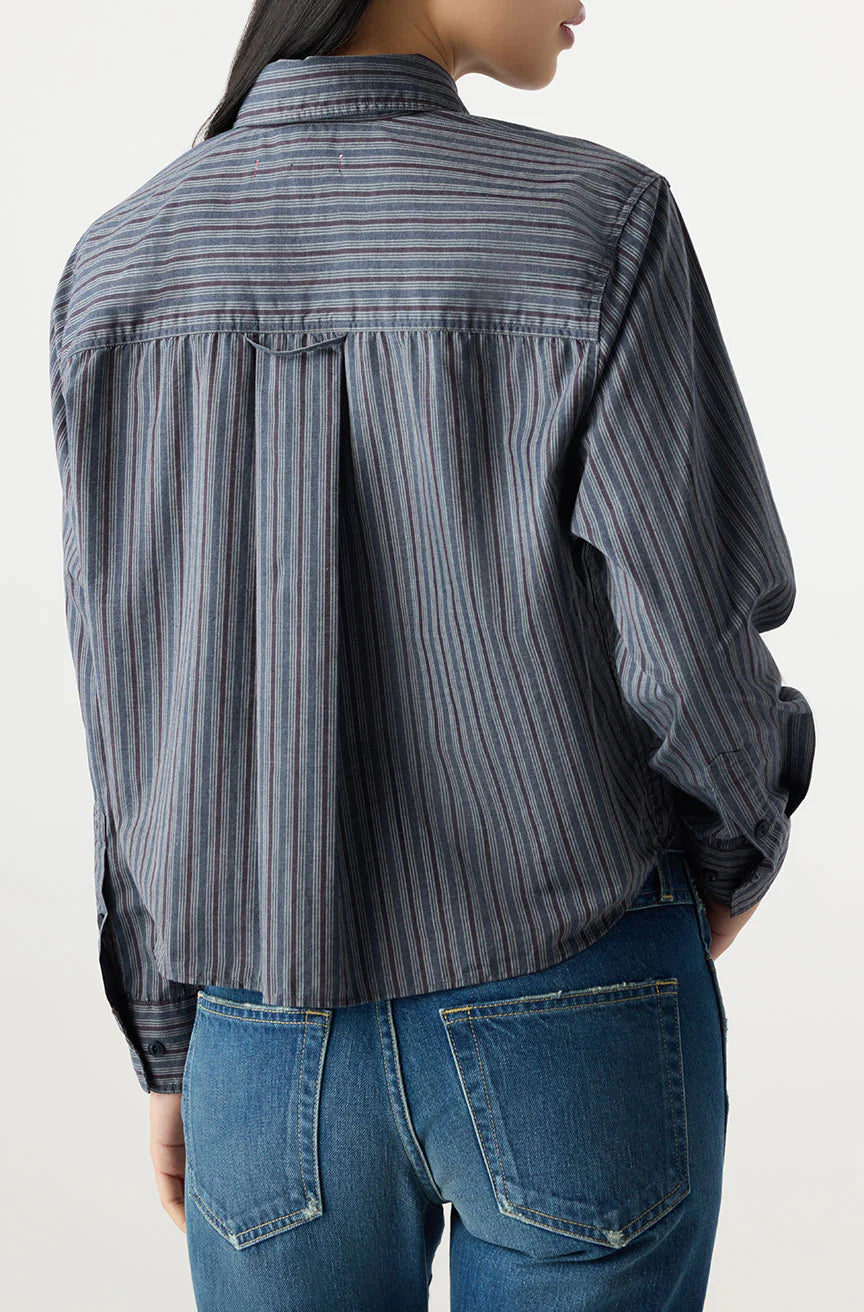 A person is shown from the back with one hand in their pocket, wearing the relaxed fit Lena Shirt by AMO Denim—a blue and gray striped button-up—paired with blue jeans against a plain light background.
