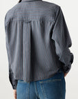 A person is shown from the back with one hand in their pocket, wearing the relaxed fit Lena Shirt by AMO Denim—a blue and gray striped button-up—paired with blue jeans against a plain light background.
