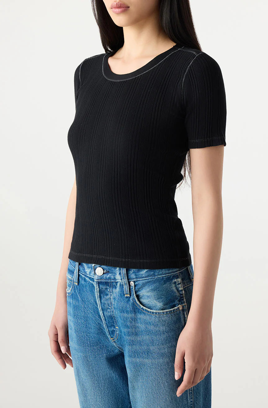 A woman wears the Cherish Tee by AMO Denim, a fitted black ribbed cotton short sleeve top, paired with blue jeans. She is shown waist up against a light background with long straight hair and her face partially out of frame.