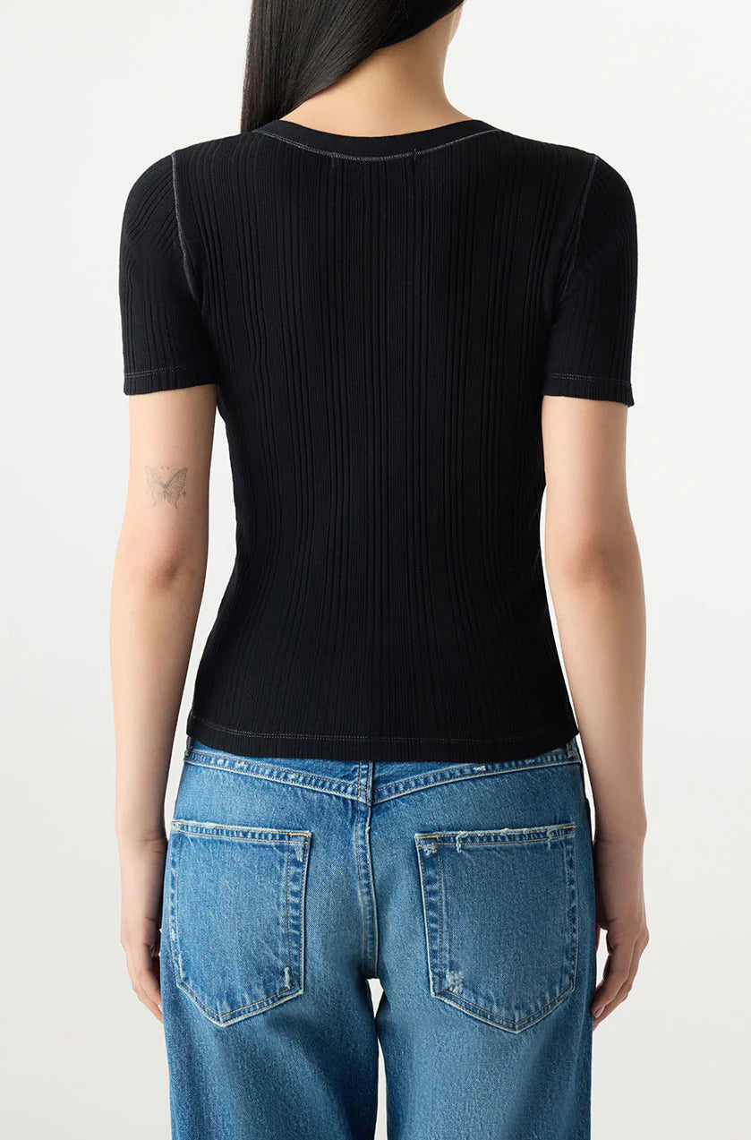 A person with long dark hair, shown from the back in blue jeans and the fitted Cherish Tee by AMO Denim, displays a small butterfly tattoo on their left arm. The ribbed cotton top features short sleeves.