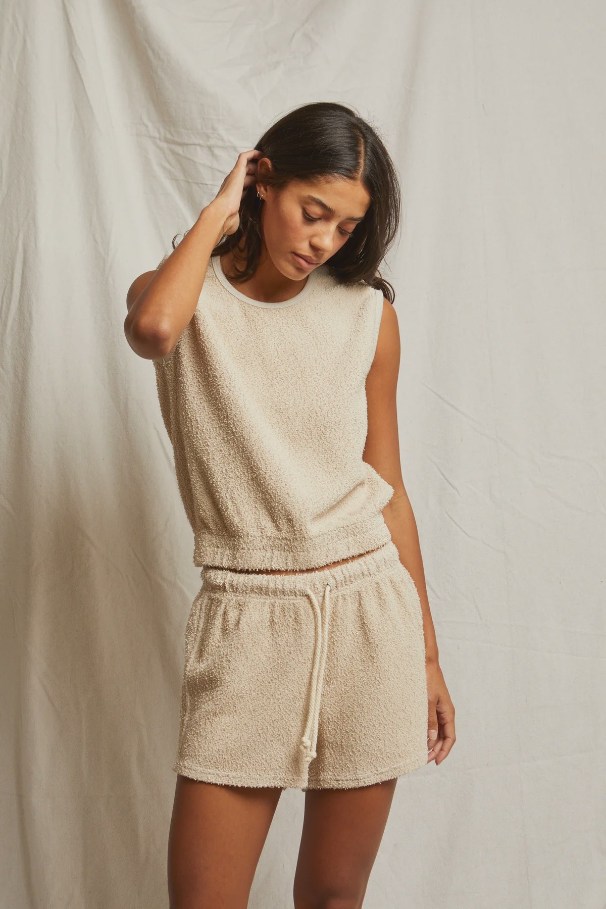 A woman stands against a light fabric backdrop, wearing the Perfectwhitetee Carli Muscle Tank and matching drawstring shorts. She looks down, touching her hair, embodying a relaxed fit with subtle vintage vibes.