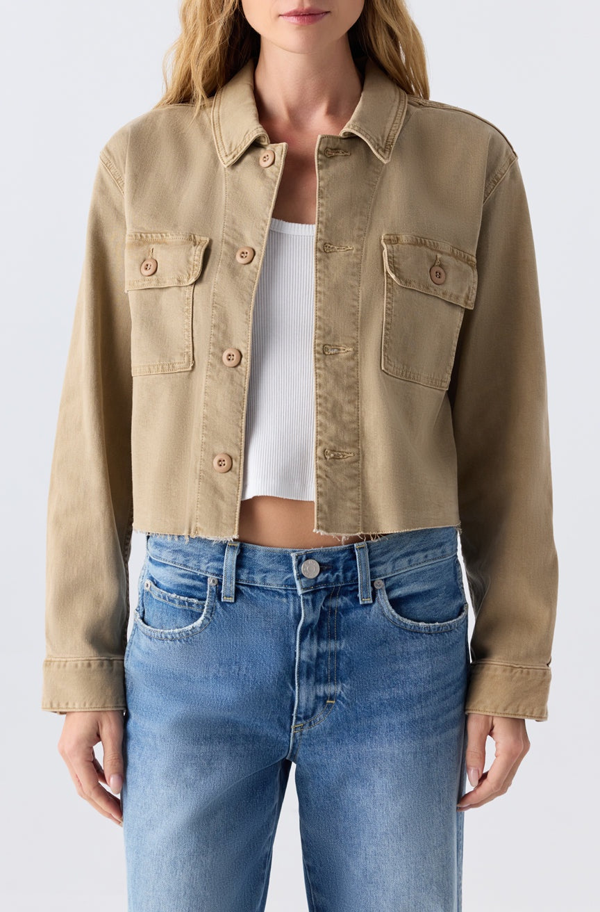 A woman wears the Carly Jacket by AMO Denim—cropped with a raw hem—over a white ribbed crop top and blue high-waisted jeans, standing against a plain light background.