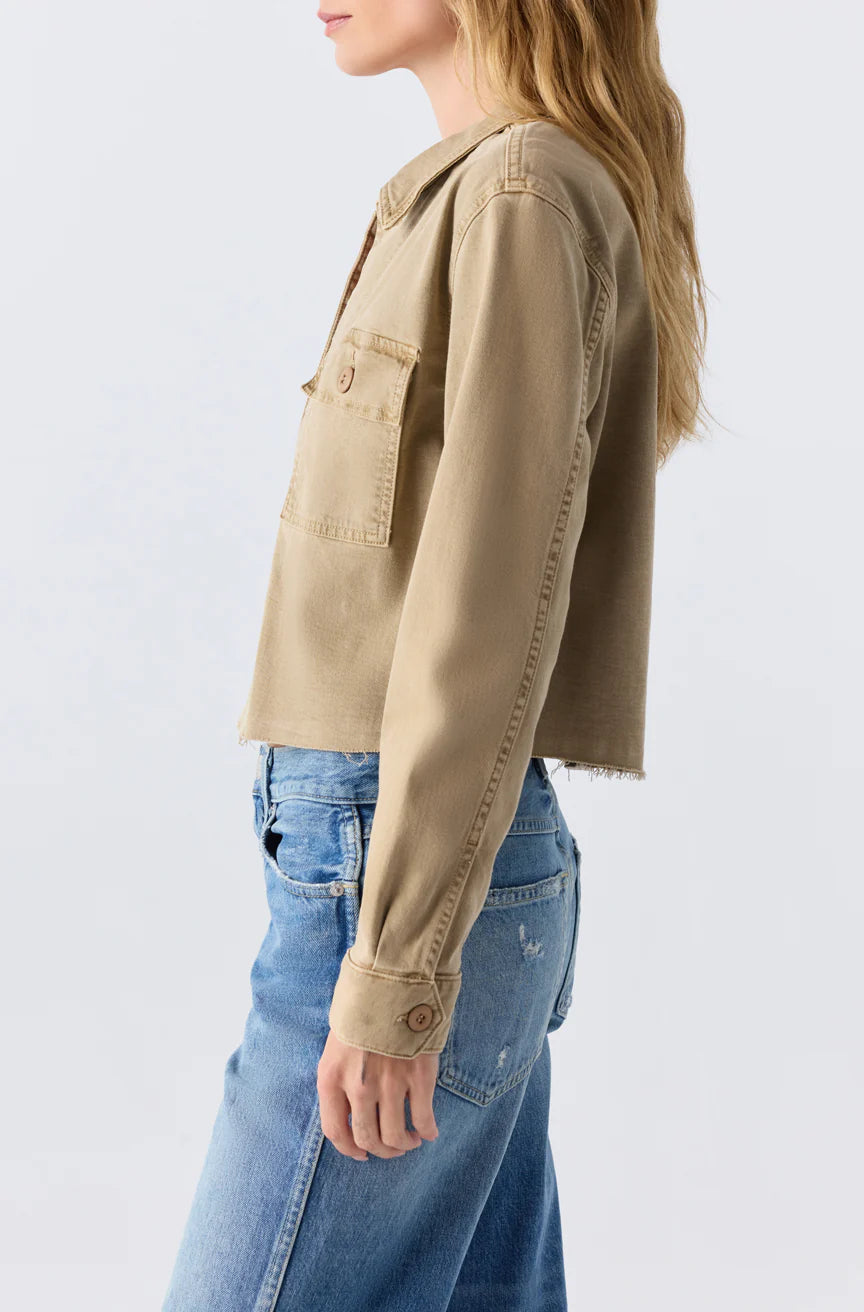 A woman stands in profile against a plain light background, wearing the AMO Denim Carly Jacket—a light brown cropped utility jacket with a chest pocket—paired with blue jeans.