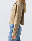 A woman stands in profile against a plain light background, wearing the AMO Denim Carly Jacket—a light brown cropped utility jacket with a chest pocket—paired with blue jeans.