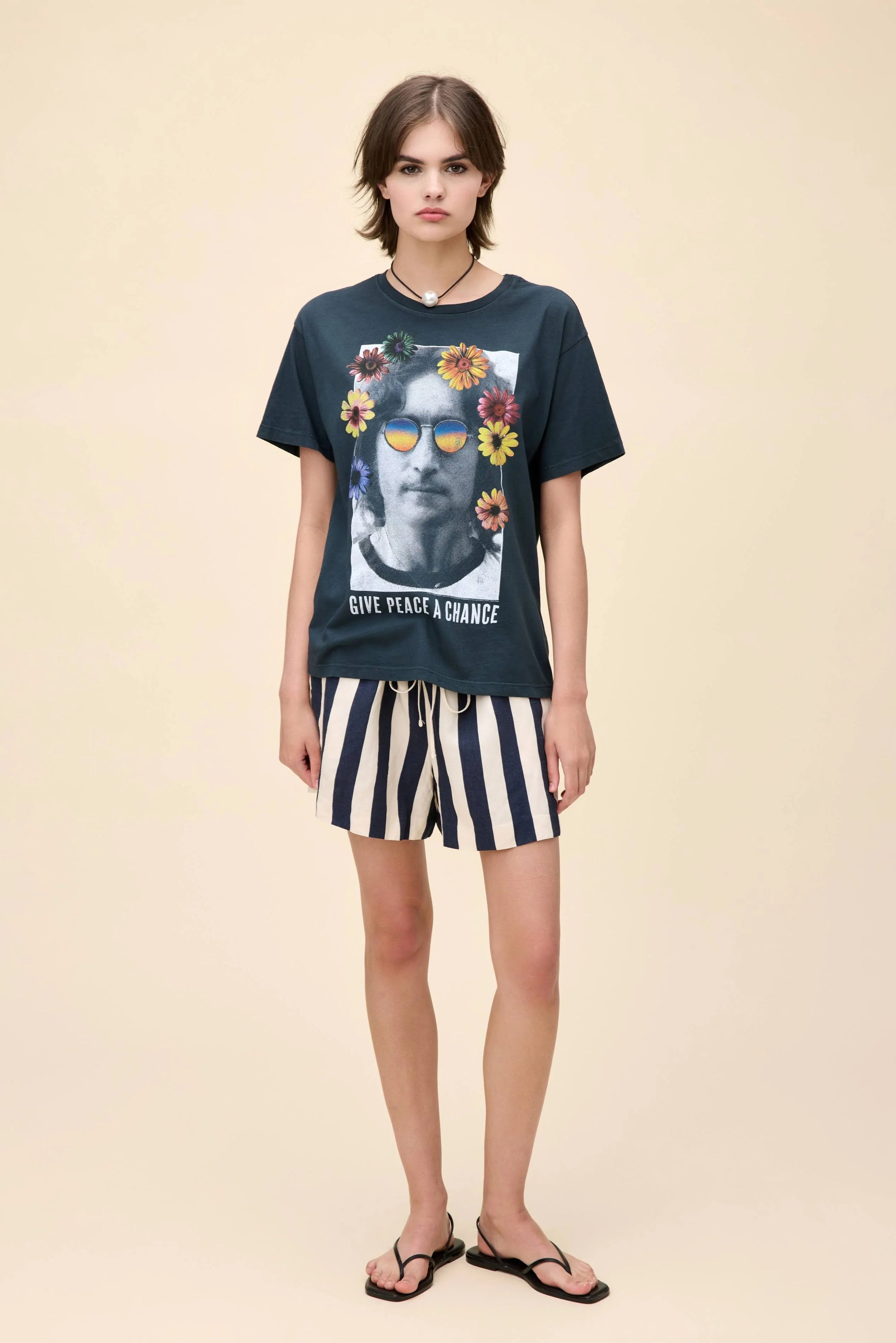 A young person stands against a beige background wearing the DayDreamer John Lennon Peace lightweight cotton black tee featuring a graphic of John Lennon's face, sunglasses, and flowers, paired with black and white striped shorts and black flip-flops.