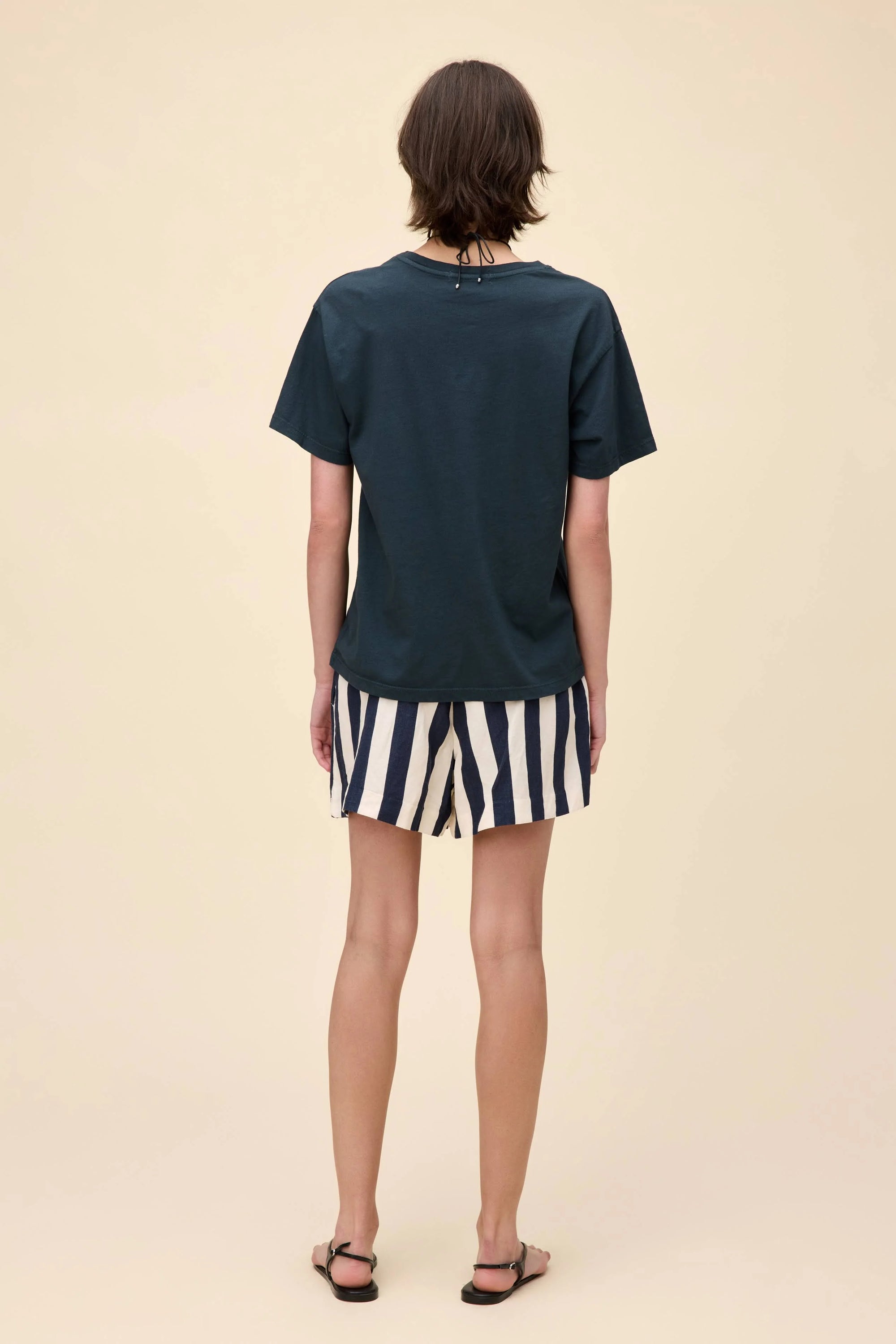 A person with short brown hair stands facing away against a plain beige background, wearing the DayDreamer John Lennon Peace Boyfriend Tee in lightweight cotton, black and white striped shorts, and black sandals.